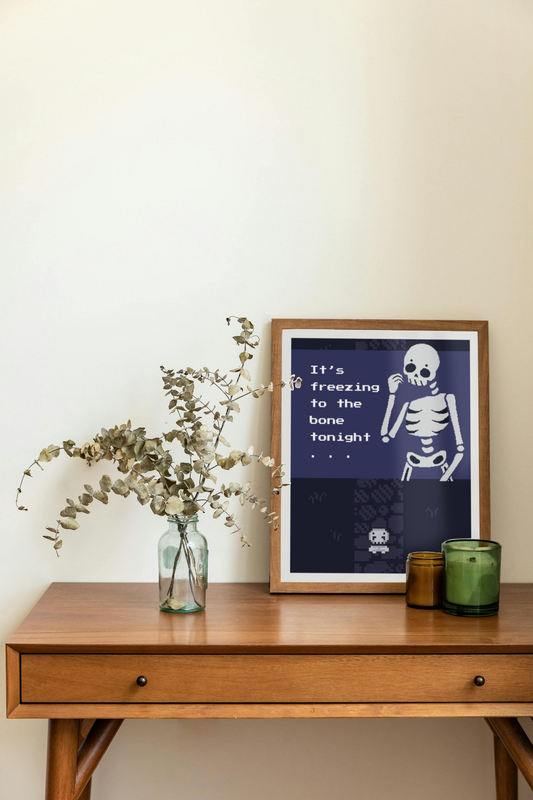 Framed art print on a desk next to a house plant and 2 candles. The framed art print is of an illustration of a pixelated skeleton with text 'It's freezing to the bone tonight' on a dark blue background. Behind that is a pixelated dark purple path on pixelated dark blue grass, with a smaller pixelated skeleton on top of that.