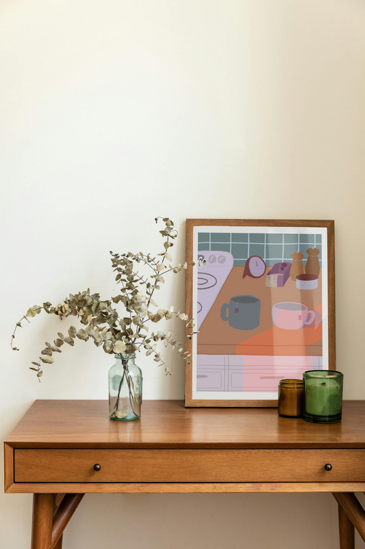 Framed art print on a desk next to a house plant and 2 candles. The framed art print is of an illustration of a kitchen counter with two mugs holding tea bags in cool evening lighting. In the background are two pepper grinders, a tea box, a salt dish, and a timer. On the left is a white stovetop, white cabinet drawers at the bottom, and green tiles for the backsplash.