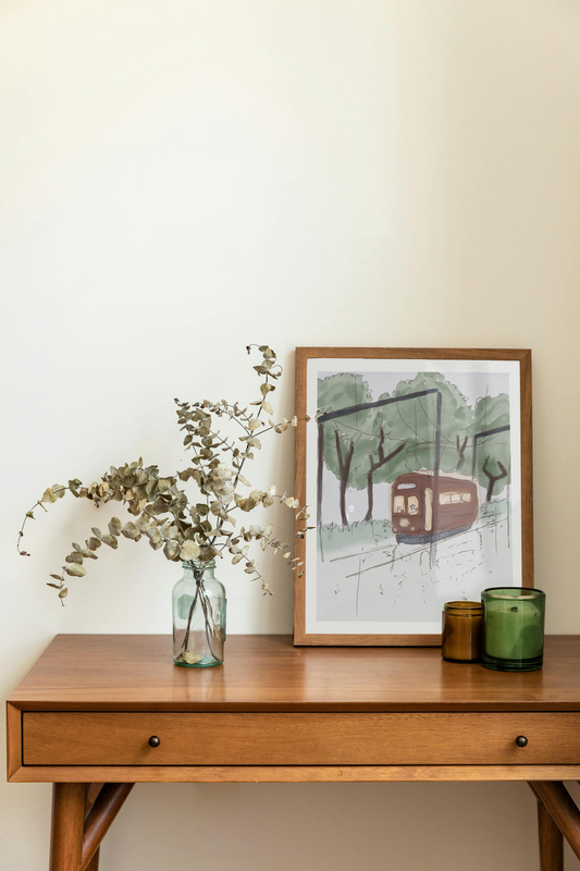 Framed art print on a desk next to a house plant and 2 candles. The framed art print is of an illustration of a watercolor-style tram in a forest with trees, a cloudy sky, and light rain. A beaver and raccoon drive the tram, while a skeleton hidden in the trees watches as it passes by.