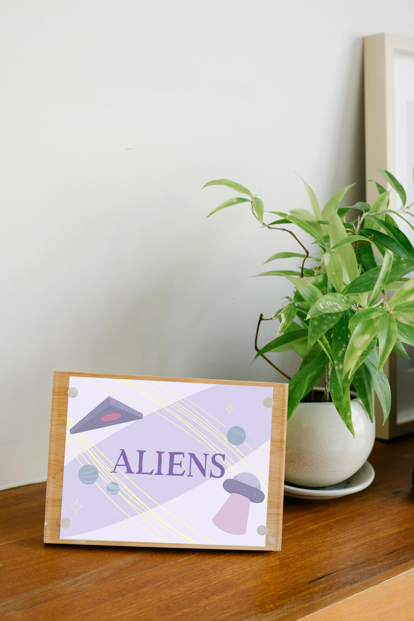 Framed art print on a wooden surface next to a small house plant of an iIllustration of a triangle UFO, a circular UFO, three green planets, and various yellow stars on a light purple background with the word 'ALIENS' in the center in dark purple.