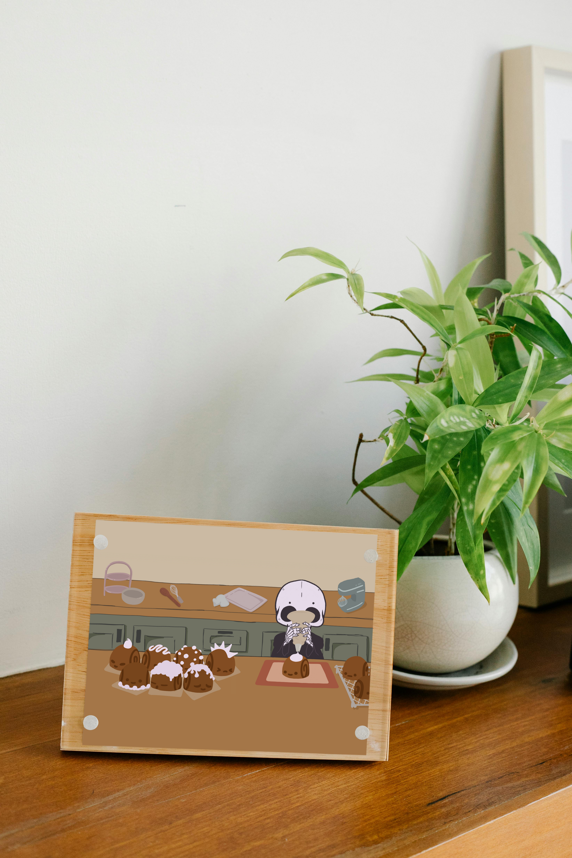 Framed art print on a wooden surface next to a small house plant of an iIllustration of a skeleton character frosting cinnamon rolls that are personified with various facial expressions and frosting designs. The skeleton stands between two wooden counters. The background consists of various kitchen supplies including a stand mixer, whisk, baking sheet, bowl, green cabinets, and a beige wall. 