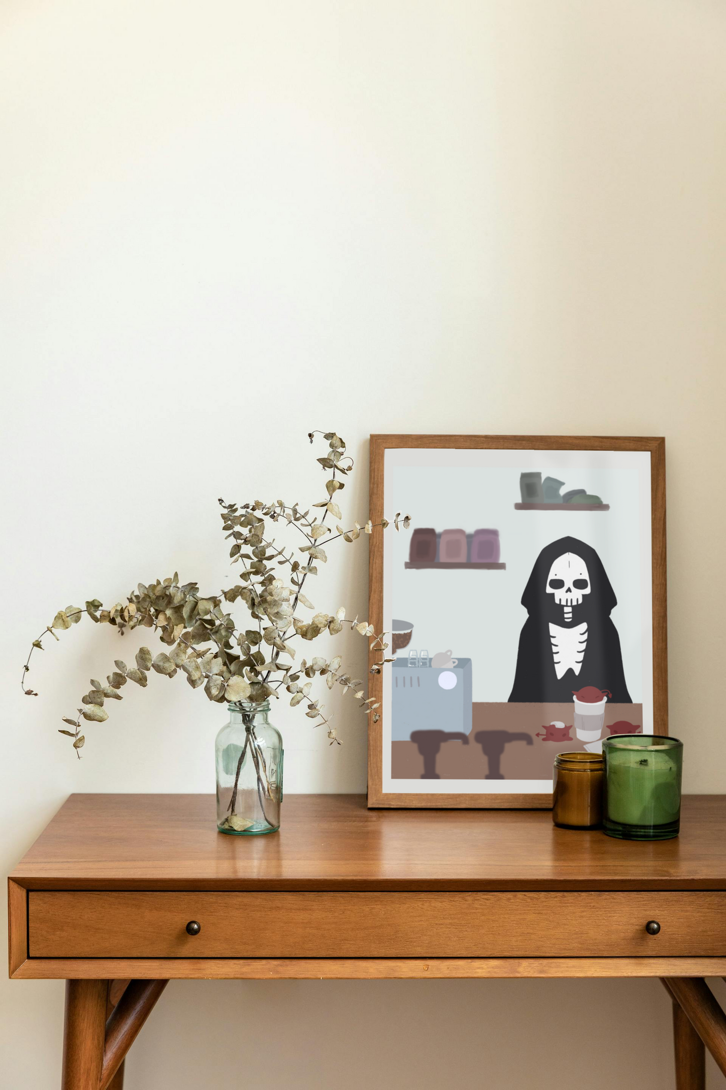 Framed art print on a desk next to a house plant and 2 candles. The framed art print is of an illustration of a skeleton character standing behind a wooden counter with an espresso machine, a to-go coffee cup, and 3 red imps atop it.  In the forefront are the tops of syrup bottles, in the background are 2 shelves holding coffee bags on a light blue wall.
