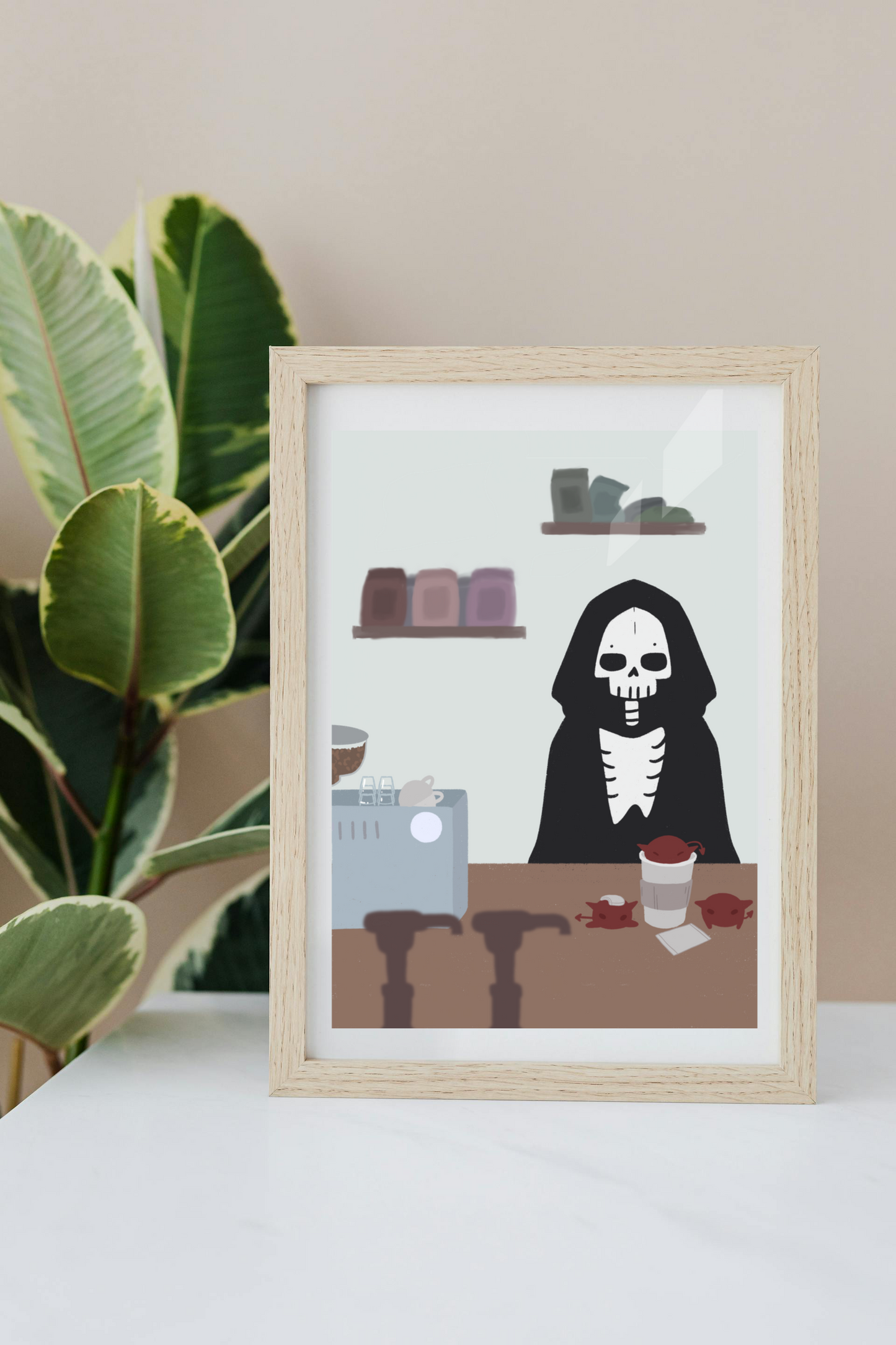 Framed art print on a white surface with a plant in the background of an illustration of a skeleton character standing behind a wooden counter with an espresso machine, a to-go coffee cup, and 3 red imps atop it.  In the forefront are the tops of syrup bottles, in the background are 2 shelves holding coffee bags on a light blue wall.