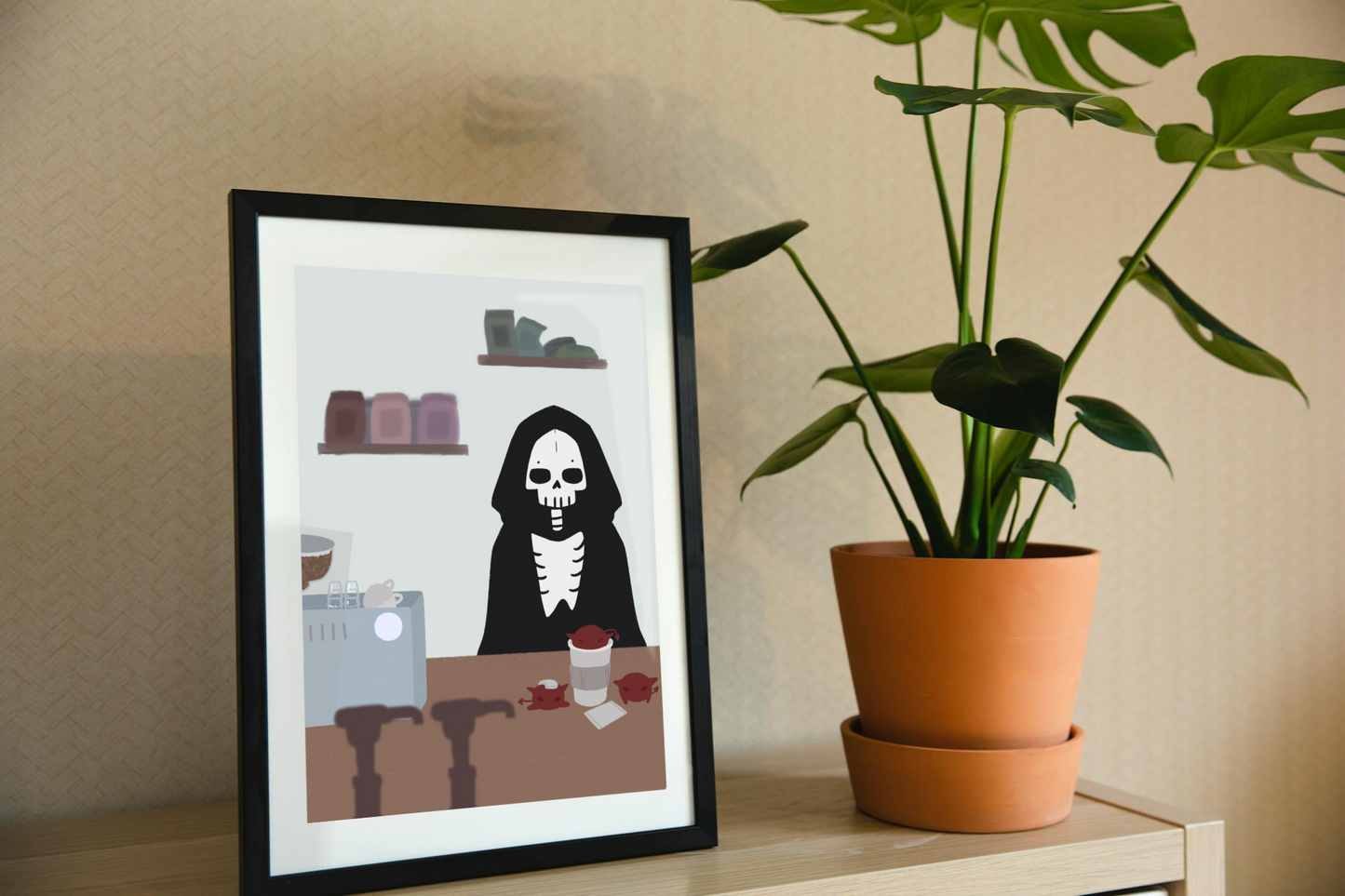 Framed art print on a shelf next to a house plant. The framed art print is of an illustration of a skeleton character standing behind a wooden counter with an espresso machine, a to-go coffee cup, and 3 red imps atop it.  In the forefront are the tops of syrup bottles, in the background are 2 shelves holding coffee bags on a light blue wall.