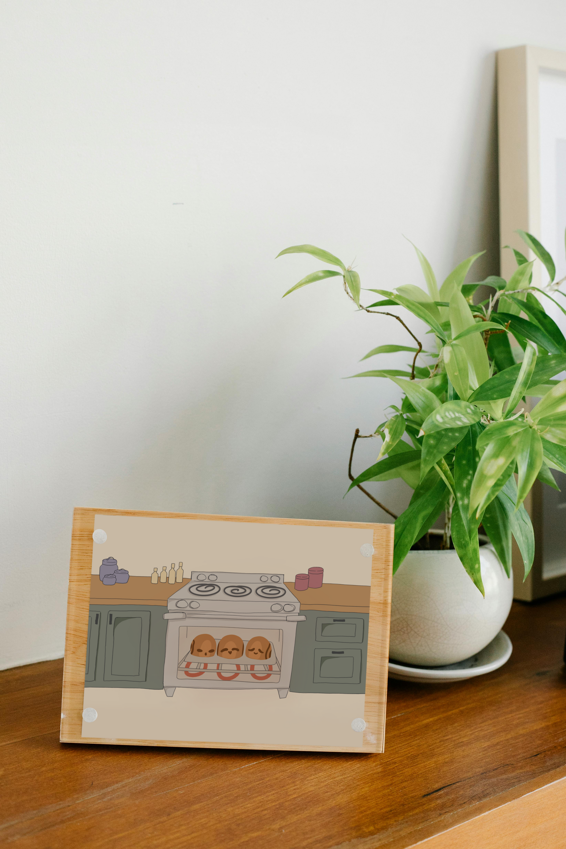 Framed art print on a wooden surface next to a small house plant of an illustration of three cinnamon rolls personified with various facial expressions in an oven surrounded by wooden kitchen counters above green cabinets in front of a beige background.