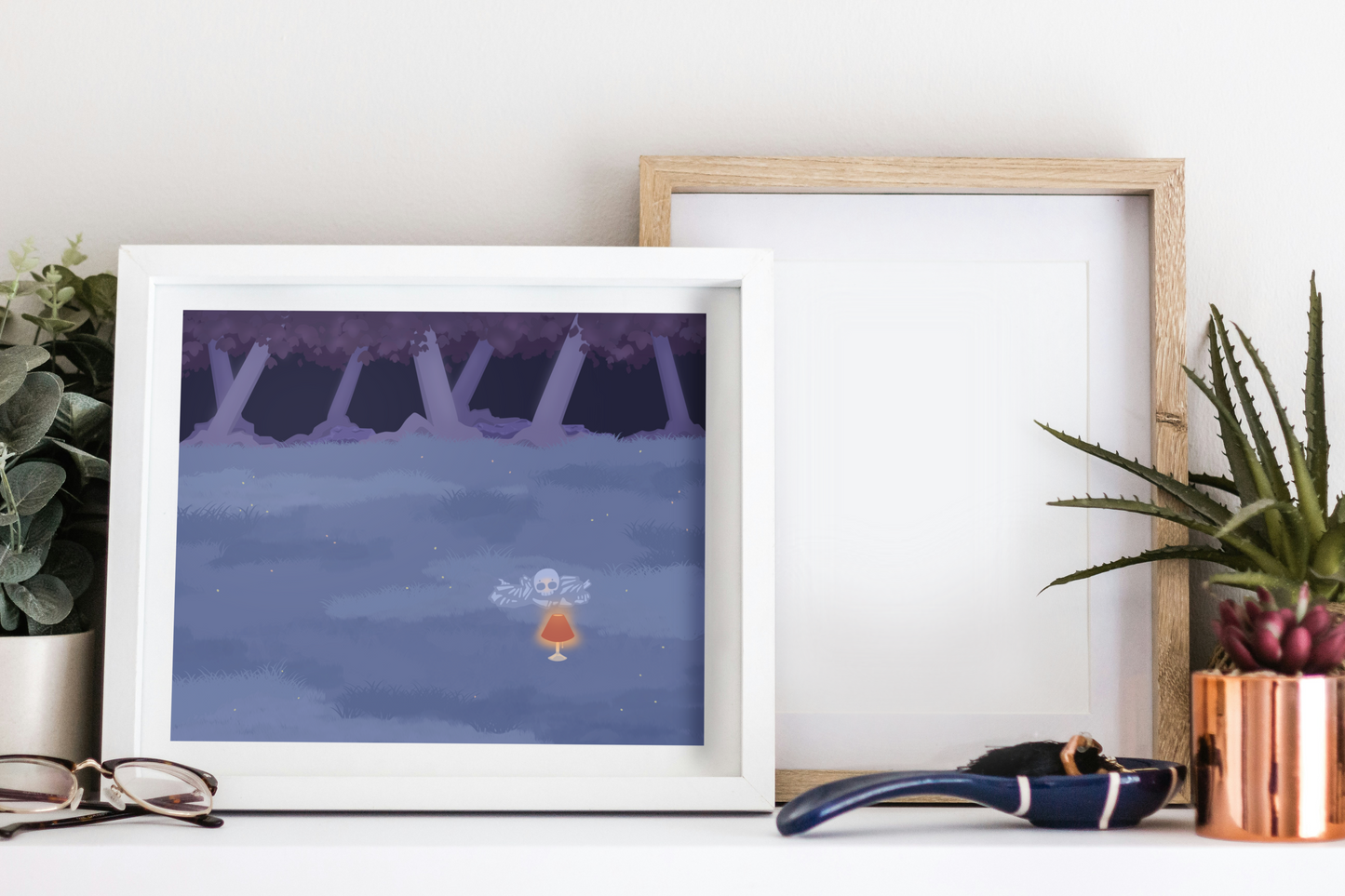Framed art print on a white surface next to small desk plants, reading glasses, and a small decorative spoon. The framed art print is of an illustration of a pile of bones in a field behind a lit lamp in the cool lighting of night. In the background is a dark forest covering the horizon.