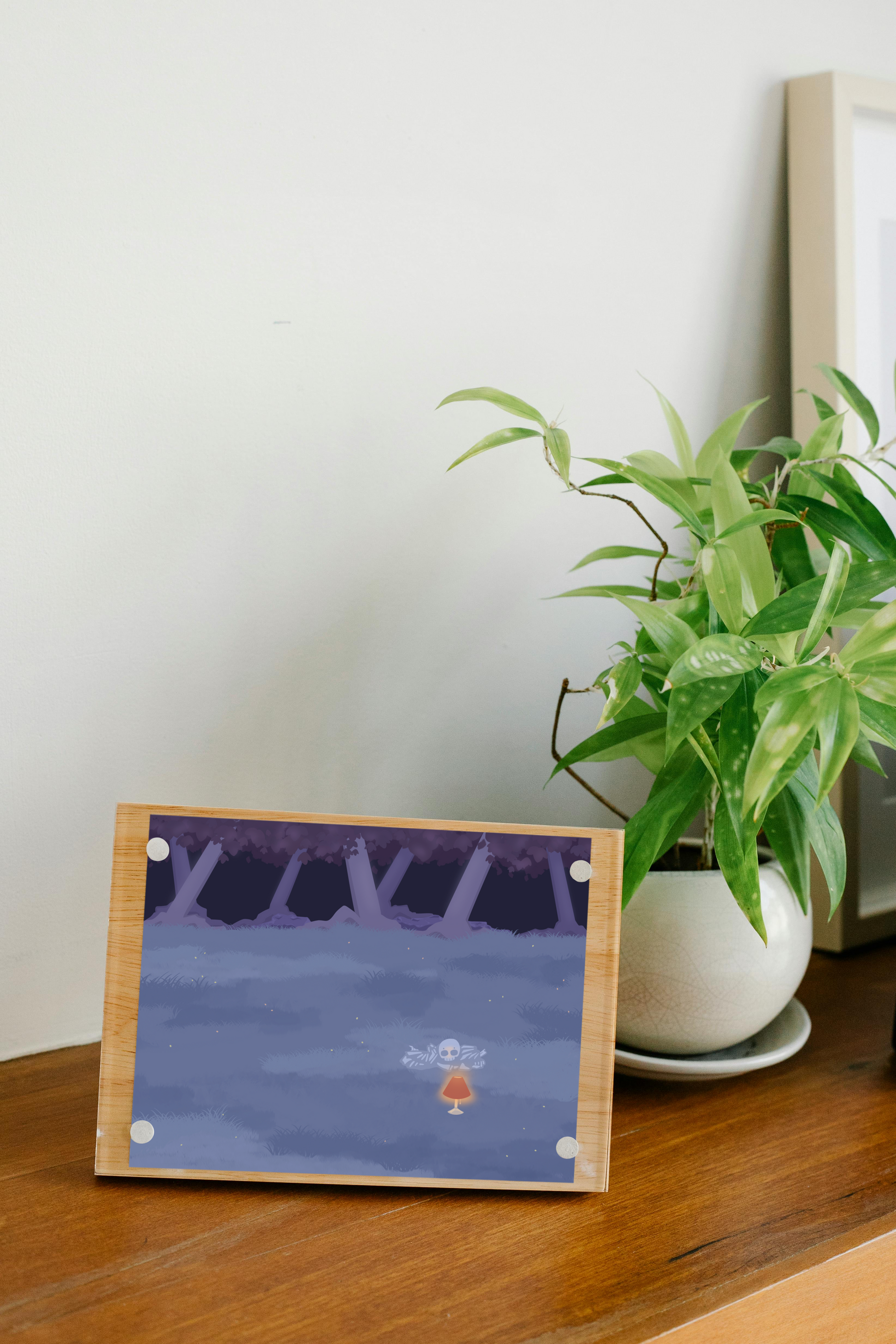 Framed art print on a wooden surface next to a small house plant of an illustration of a pile of bones in a field behind a lit lamp in the cool lighting of night. In the background is a dark forest covering the horizon.