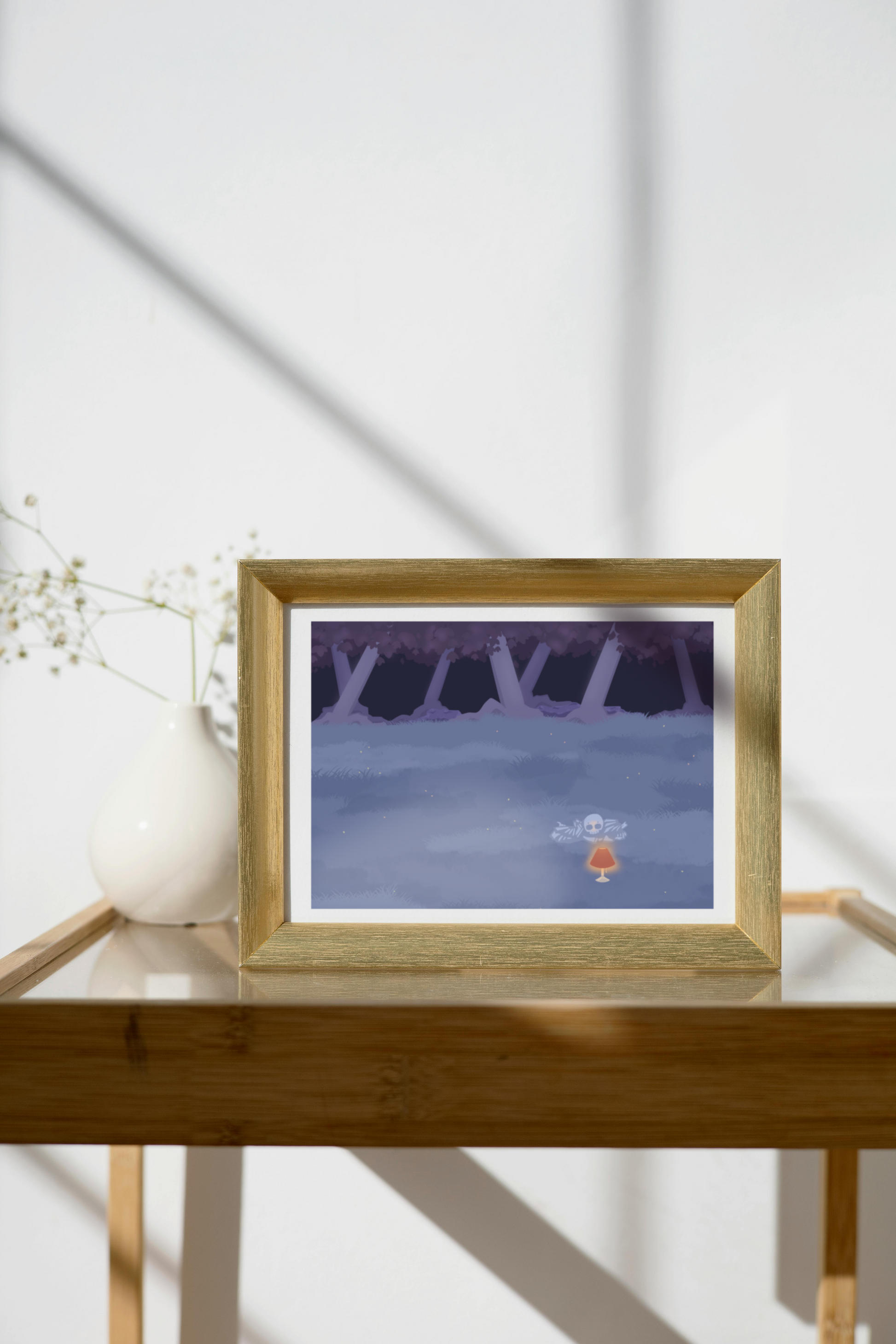 Framed art print on a glass and wooden side table next to a small plant in a white, ceramic vase. The framed art print is of an illustration of a pile of bones in a field behind a lit lamp in the cool lighting of night. In the background is a dark forest covering the horizon.