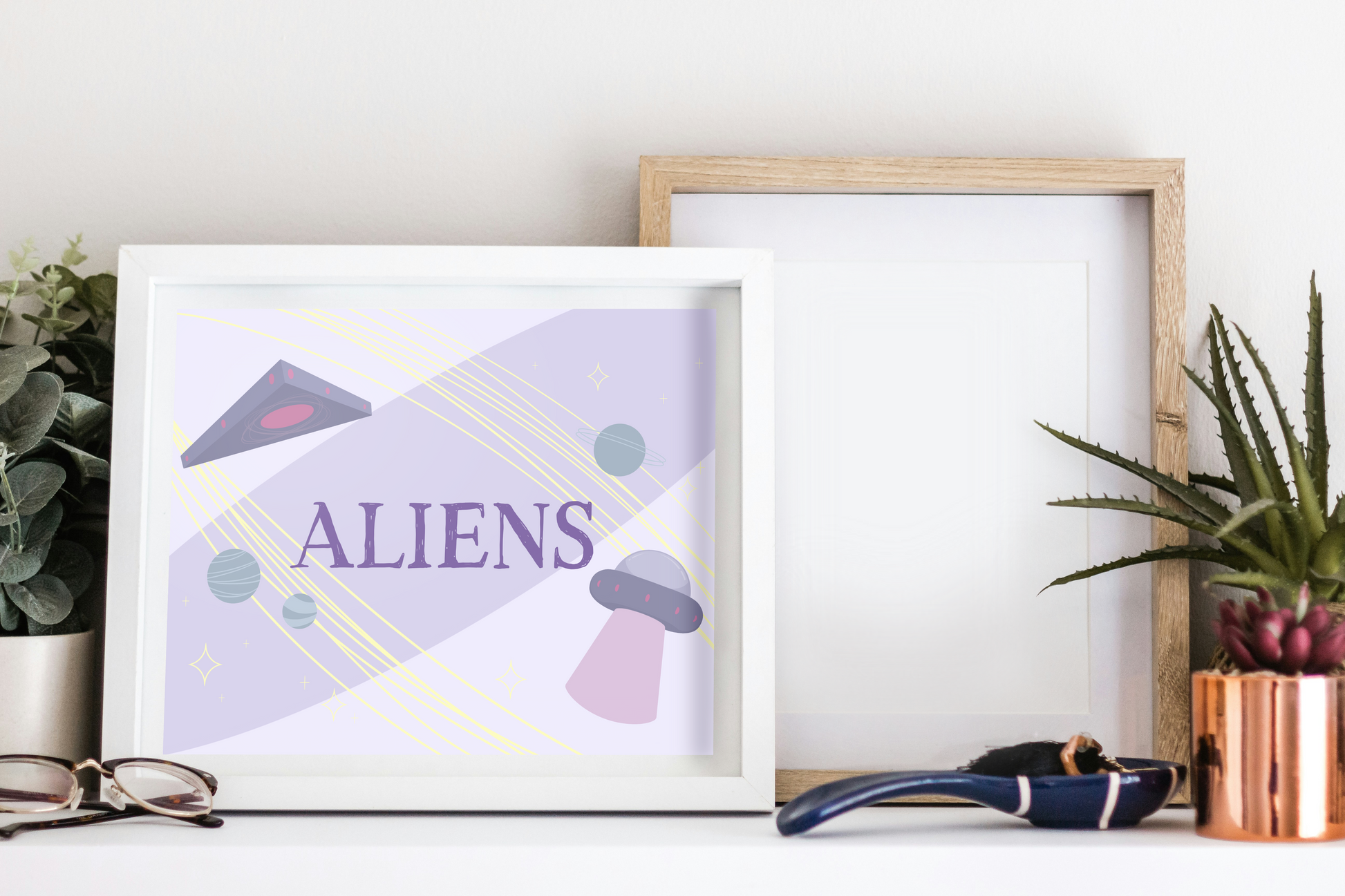 Framed art print on a white surface next to small desk plants, reading glasses, and a small decorative spoon. The framed art print is of an illustration of a triangle UFO, a circular UFO, three green planets, and various yellow stars on a light purple background with the word 'ALIENS' in the center in dark purple.