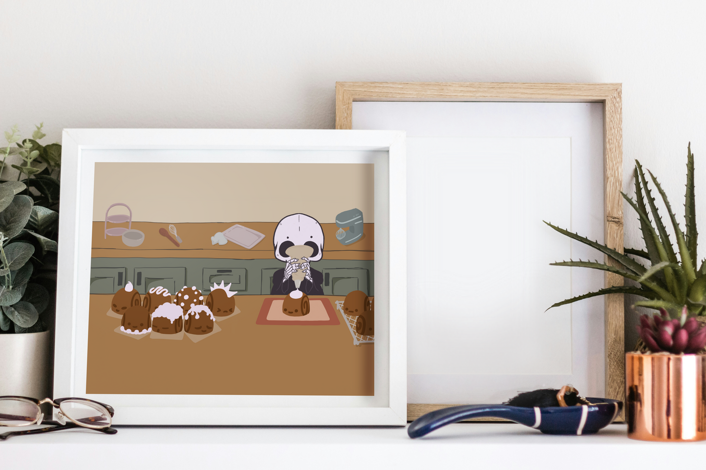 Framed art print on a white surface next to small desk plants, reading glasses, and a small decorative spoon. The framed art print is of an illustration of a skeleton character frosting cinnamon rolls that are personified with various facial expressions and frosting designs. The skeleton stands between two wooden counters. The background consists of various kitchen supplies including a stand mixer, whisk, baking sheet, bowl, green cabinets, and a beige wall. 