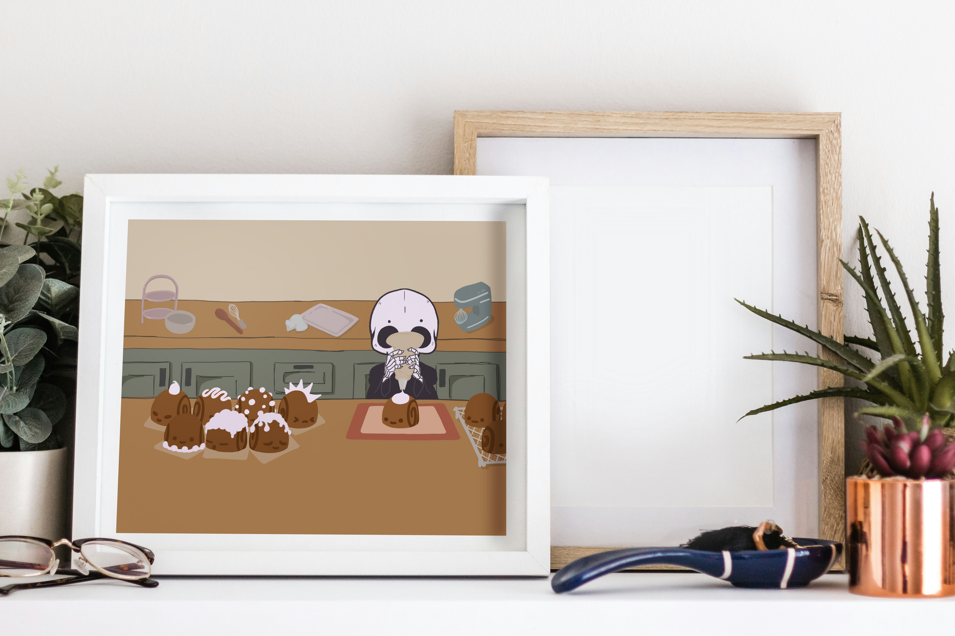 Framed art print on a white surface next to small desk plants, reading glasses, and a small decorative spoon. The framed art print is of an illustration of a skeleton character frosting cinnamon rolls that are personified with various facial expressions and frosting designs. The skeleton stands between two wooden counters. The background consists of various kitchen supplies including a stand mixer, whisk, baking sheet, bowl, green cabinets, and a beige wall. 