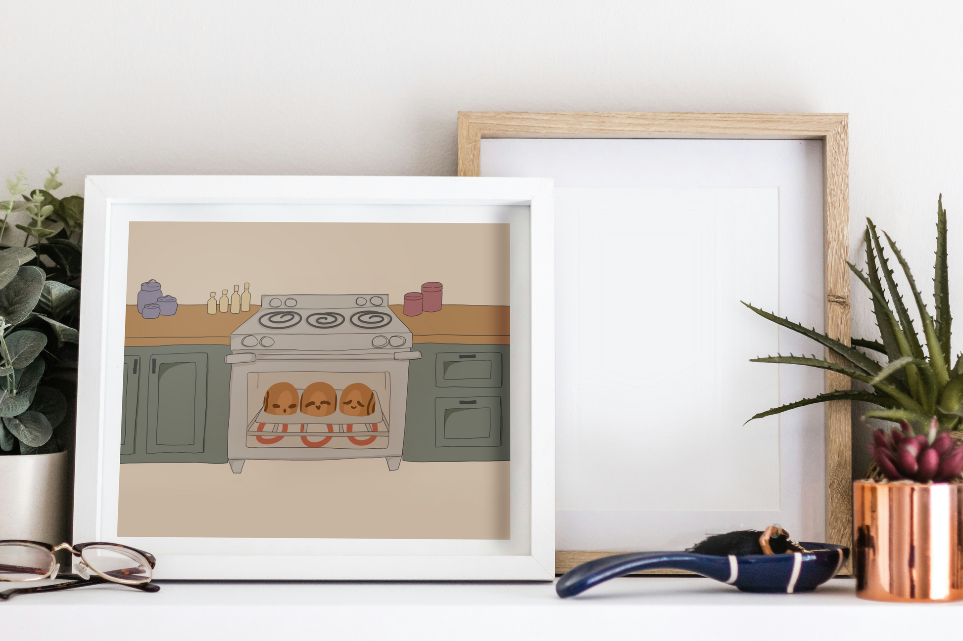 Framed art print on a white surface next to small desk plants, reading glasses, and a small decorative spoon. The framed art print is of an illustration of three cinnamon rolls personified with various facial expressions in an oven surrounded by wooden kitchen counters above green cabinets in front of a beige background.