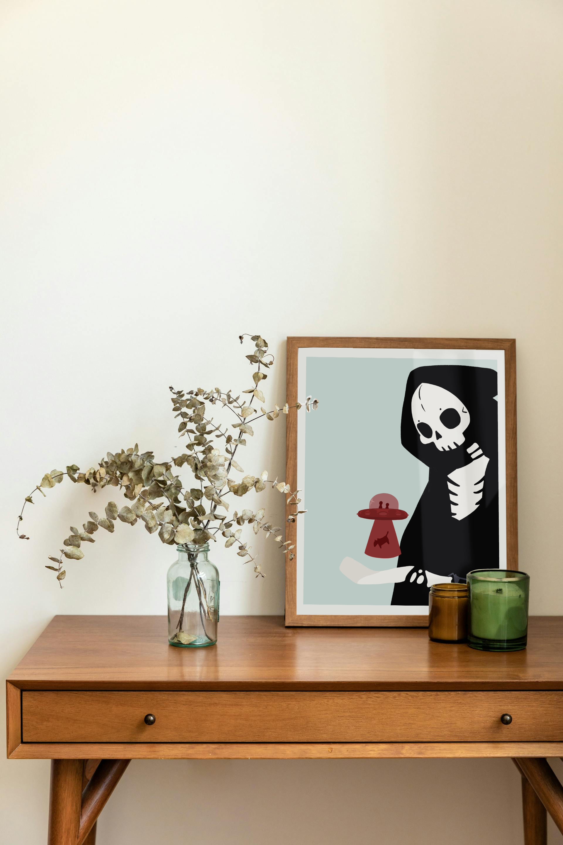 Framed art print on a desk next to a house plant and 2 candles. The framed art print is of an illustration of a skeletal character with a red UFO abducting a cow hovering over its outreached hand in front of a light blue background.