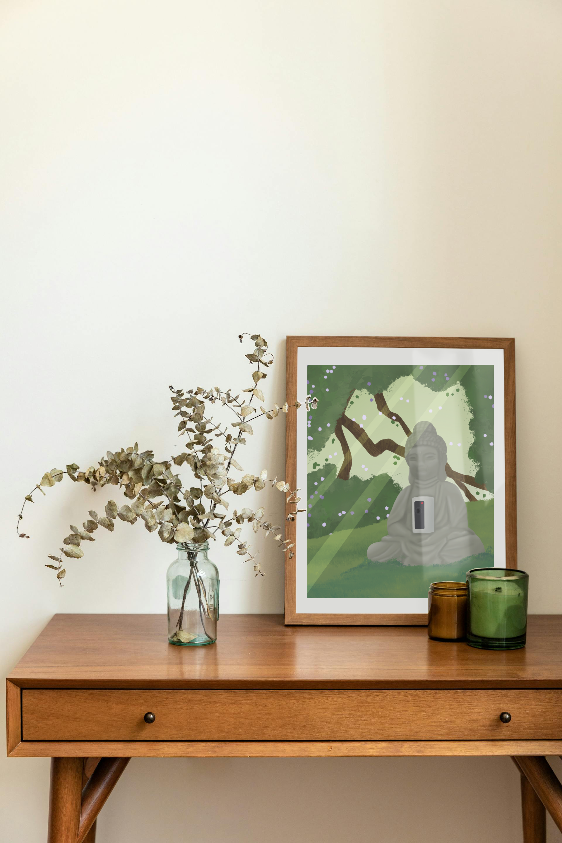 Framed art print on a desk next to a house plant and 2 candles. The framed art print is of an illustration of a buddha statue with a security camera embedded into it, surrounded by trees, grass, and sunlight.