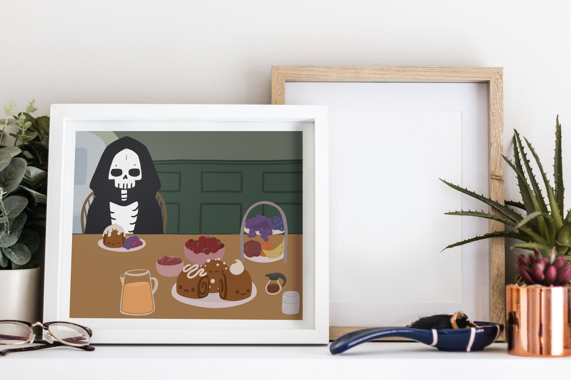 Framed art print on a white surface next to small desk plants, reading glasses, and a small decorative spoon. The framed art print is of an illustration of a skeleton character sitting at a table with brunch foods, including orange juice, cinnamon rolls personified with faces and various frosting designs, various fruits, a bowl of cherry tomatoes, syrup, jam, and powdered sugar. The background is of a green wall and hallway.