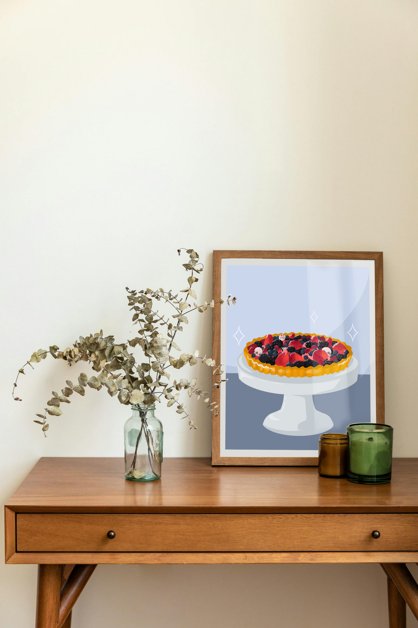 Framed art print on a desk next to a house plant and 2 candles. The framed art print is of an illustration of a fruit tart filled with strawberries, blackberries, raspberries, blueberries, tiny skulls, and tiny hand bones on a gray cake stand against a light blue and dark blue background. Three sparkles surround the fruit tart.