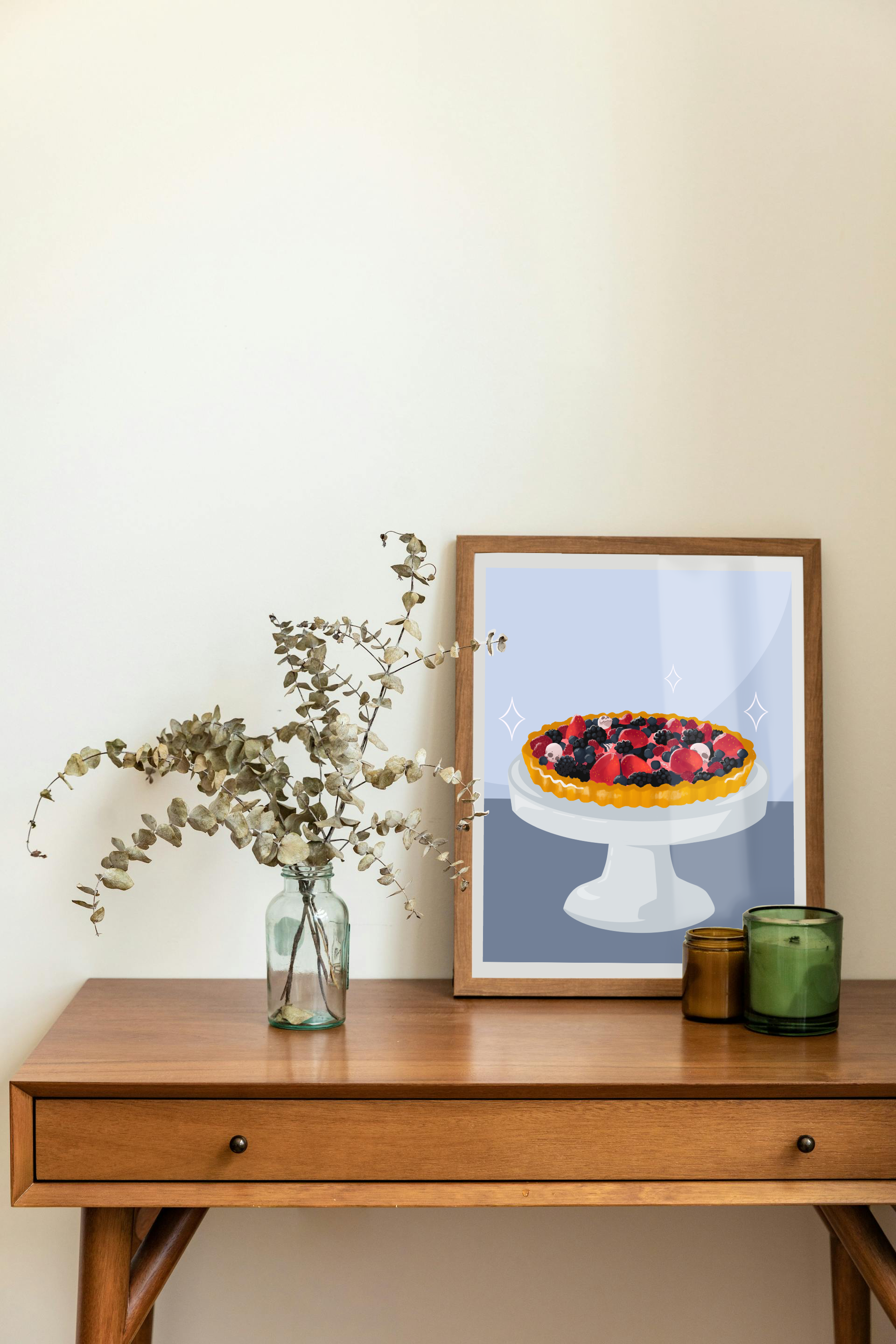 Framed art print on a desk next to a house plant and 2 candles. The framed art print is of an illustration of a fruit tart filled with strawberries, blackberries, raspberries, blueberries, tiny skulls, and tiny hand bones on a gray cake stand against a light blue and dark blue background. Three sparkles surround the fruit tart.