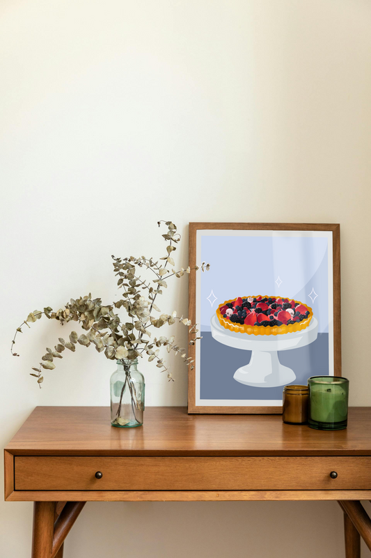 Framed art print on a desk next to a house plant and 2 candles. The framed art print is of an illustration of a fruit tart filled with strawberries, blackberries, raspberries, blueberries, tiny skulls, and tiny hand bones on a gray cake stand against a light blue and dark blue background. Three sparkles surround the fruit tart.