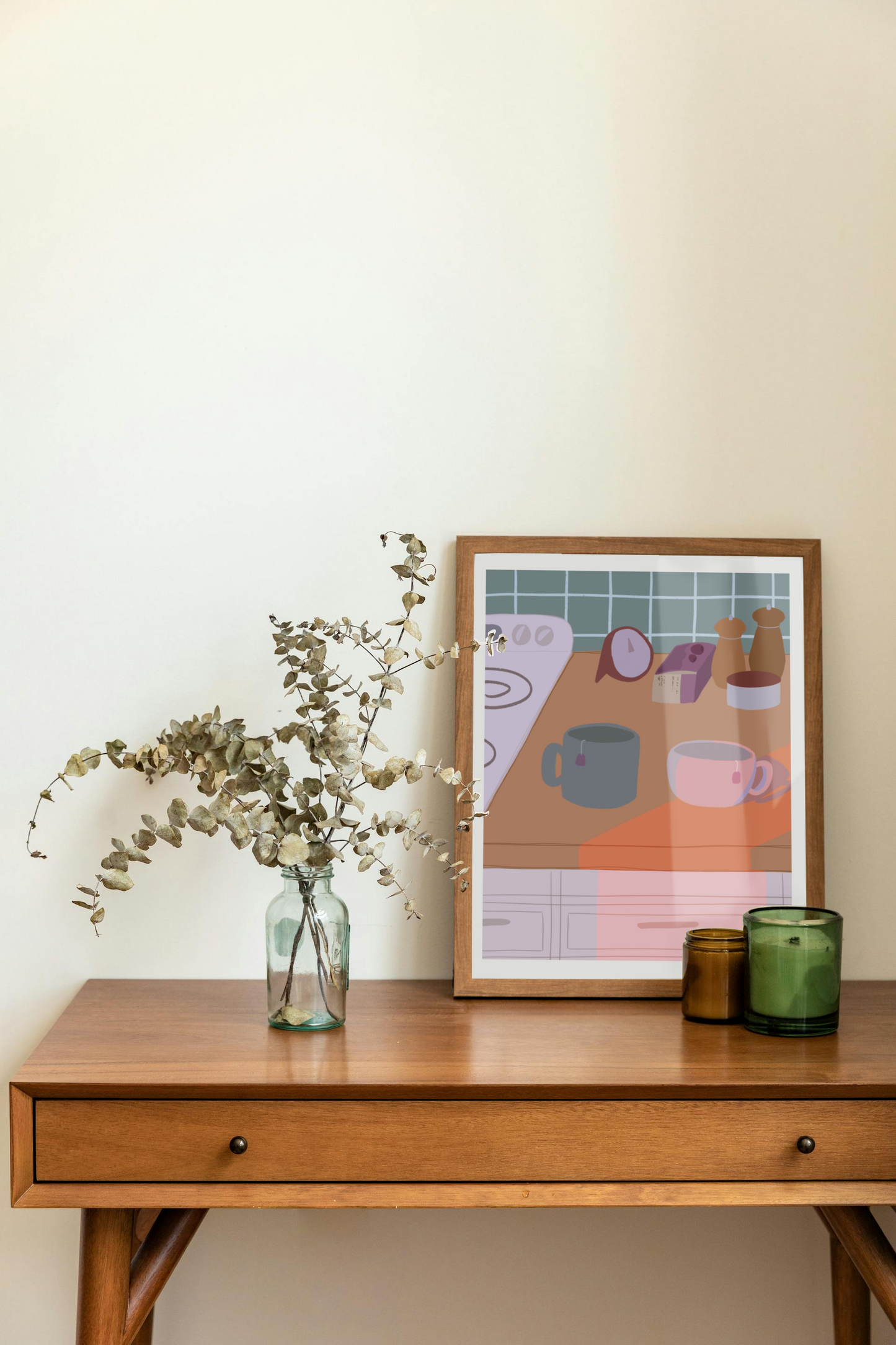Framed art print on a desk next to a house plant and 2 candles. The framed art print is of an illustration of a kitchen counter with two mugs holding tea bags in cool evening lighting. In the background are two pepper grinders, a tea box, a salt dish, and a timer. On the left is a white stovetop, white cabinet drawers at the bottom, and green tiles for the backsplash.