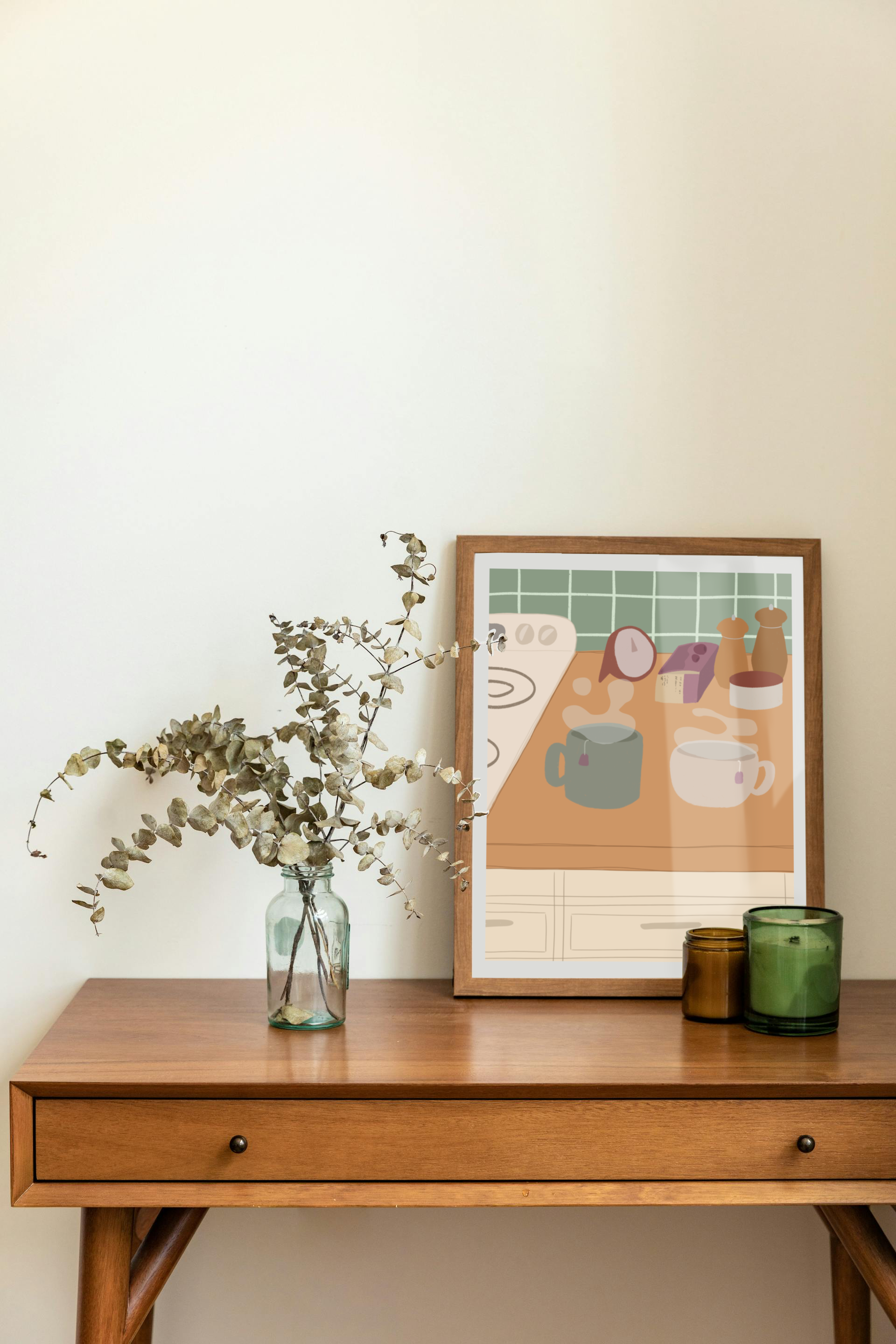 Framed art print on a desk next to a house plant and 2 candles. The framed art print is of an illustration of a kitchen counter with two steaming mugs holding tea bags in morning lighting. In the background are two pepper grinders, a tea box, a salt dish, and a timer. On the left is a white stovetop, white cabinet drawers at the bottom, and green tiles for the backsplash.