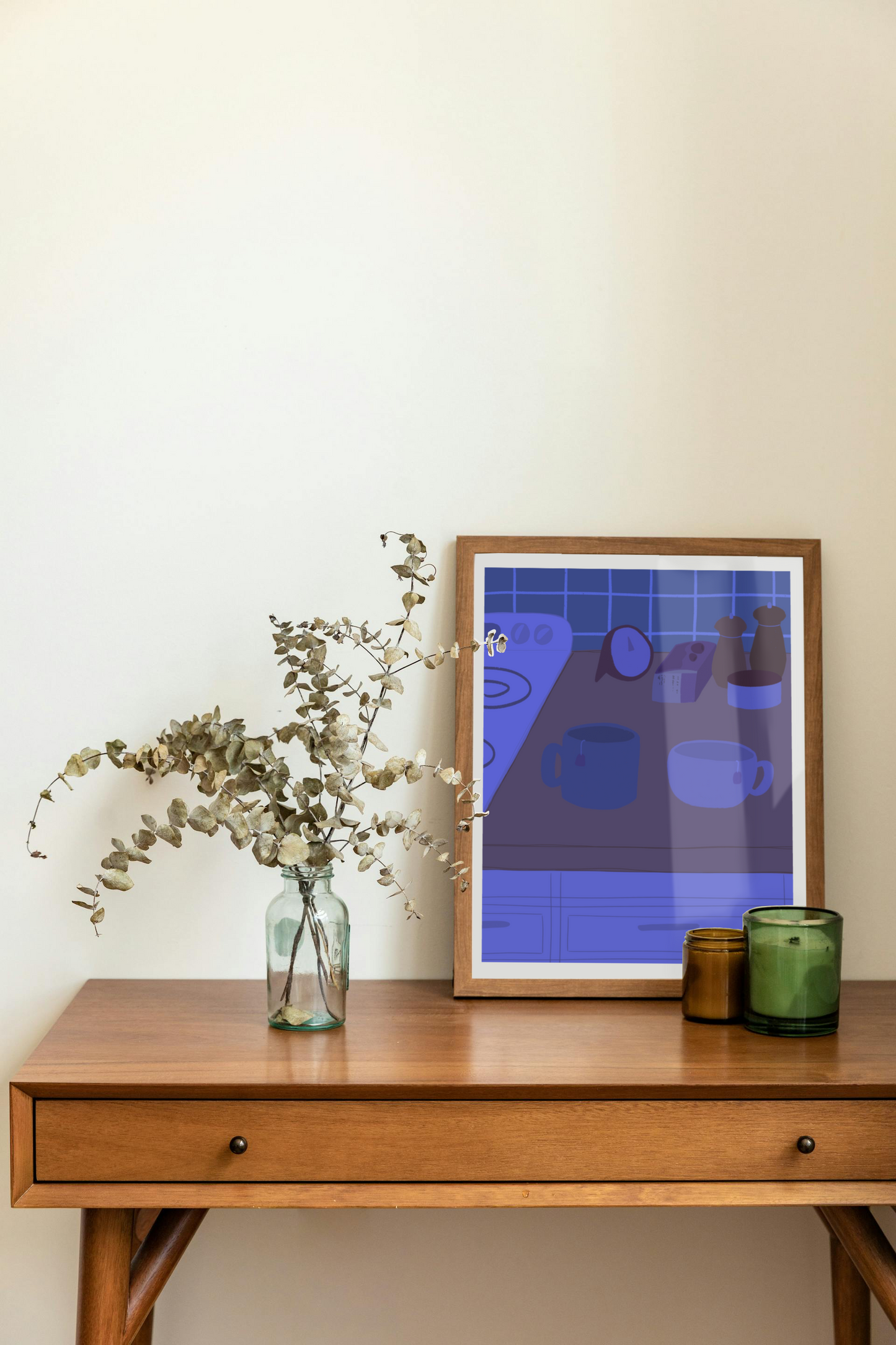 Framed art print on a desk next to a house plant and 2 candles. The framed art print is of an illustration of a kitchen counter with two mugs holding tea bags in blue night lighting. In the background are two pepper grinders, a tea box, a salt dish, and a timer. On the left is a white stovetop, white cabinet drawers at the bottom, and green tiles for the backsplash.