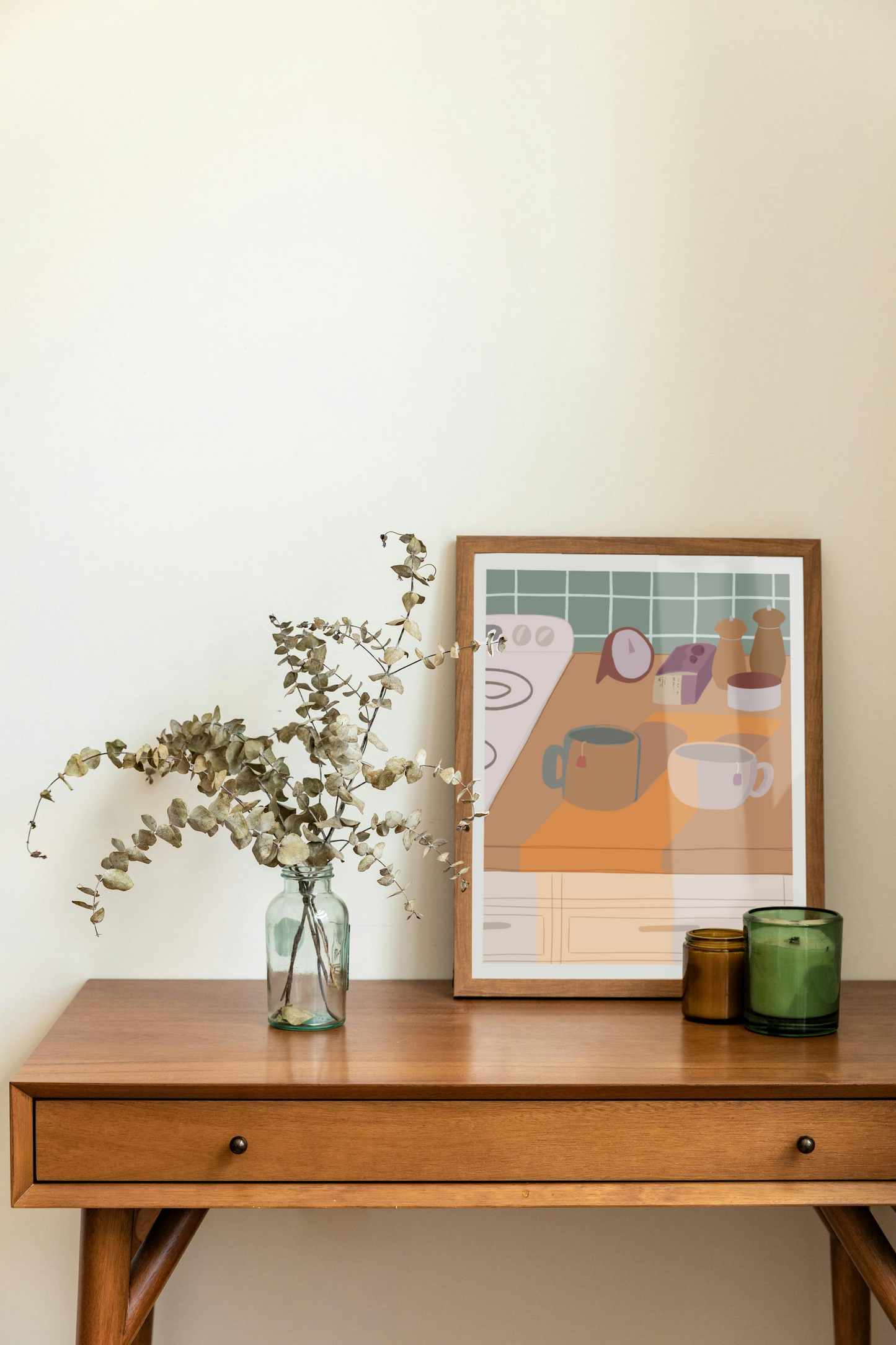 Framed art print on a desk next to a house plant and 2 candles. The framed art print is of an illustration of a kitchen counter with two mugs holding tea bags in warm afternoon lighting. In the background are two pepper grinders, a tea box, a salt dish, and a timer. On the left is a white stovetop, white cabinet drawers at the bottom, and green tiles for the backsplash.