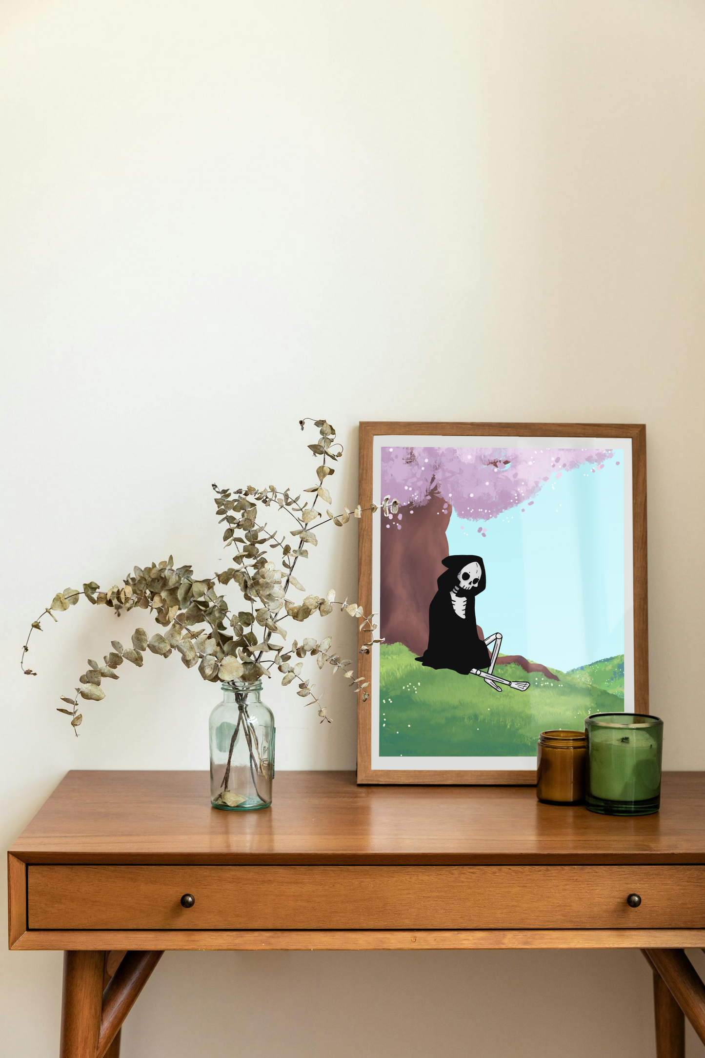 Framed art print on a desk next to a house plant and 2 candles. The framed art print is of a skeleton under a cherry blossom tree with a light blue sky in a hilly field.