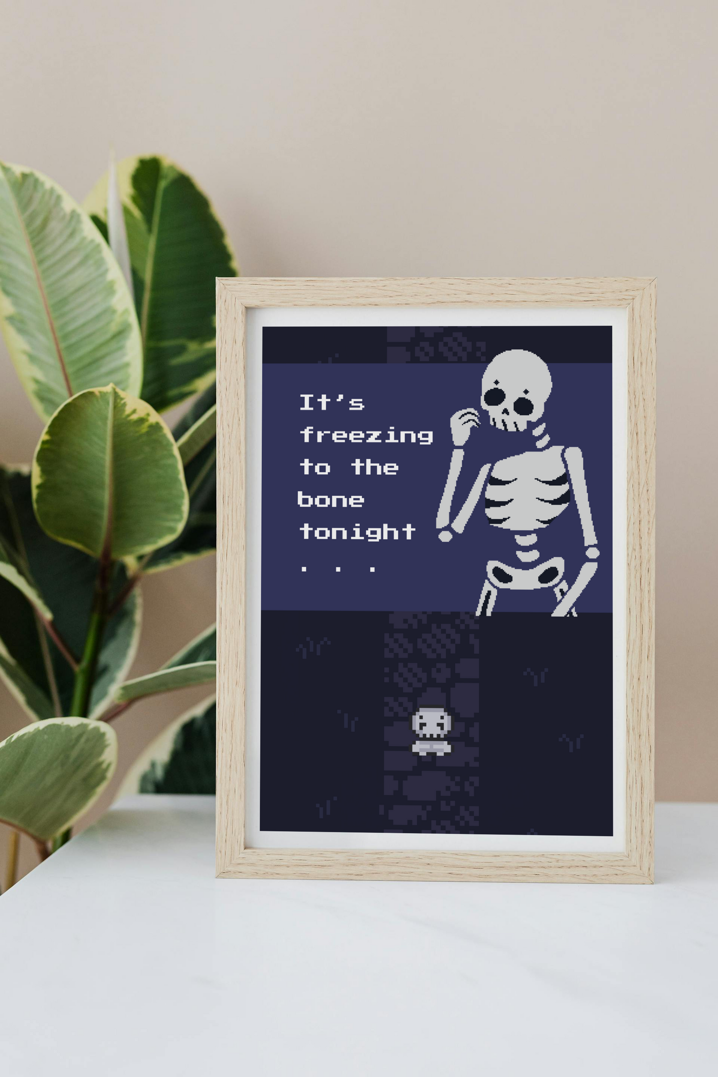 Framed art print on a white surface with a plant in the background of an illustration of a pixelated skeleton with text 'It's freezing to the bone tonight' on a dark blue background. Behind that is a pixelated dark purple path on pixelated dark blue grass, with a smaller pixelated skeleton on top of that.