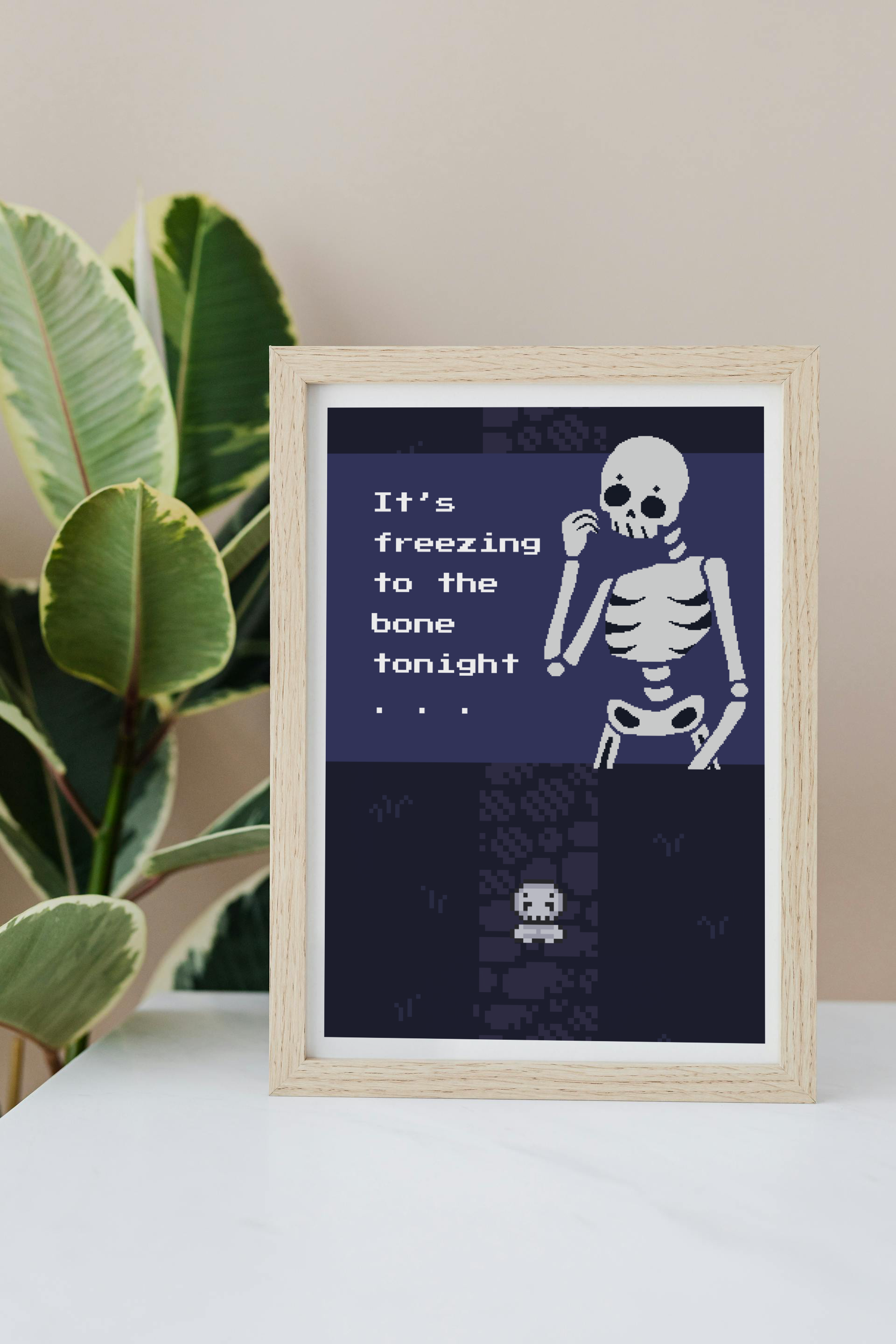 Framed art print on a white surface with a plant in the background of an illustration of a pixelated skeleton with text 'It's freezing to the bone tonight' on a dark blue background. Behind that is a pixelated dark purple path on pixelated dark blue grass, with a smaller pixelated skeleton on top of that.