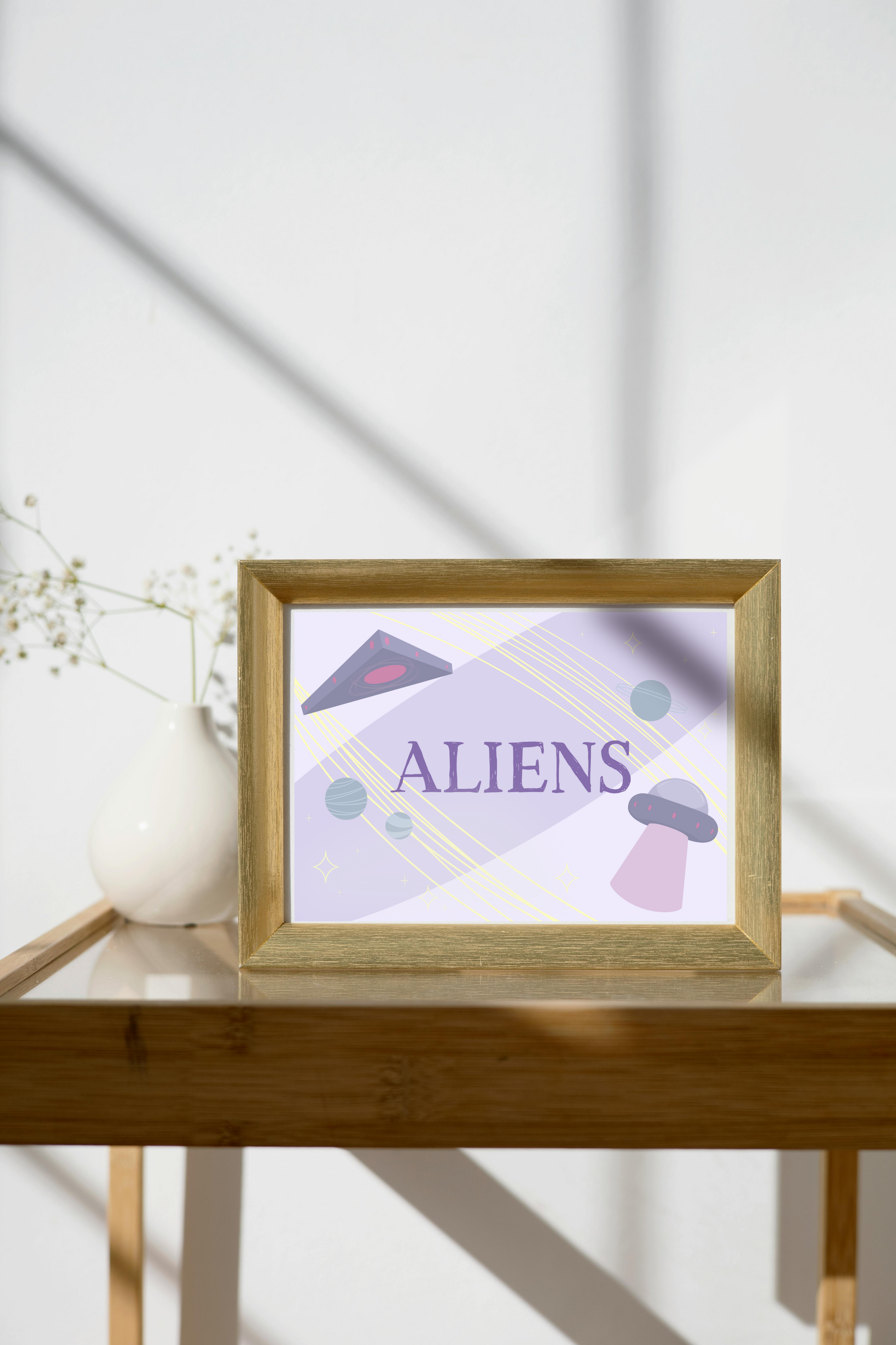 Framed art print on a glass and wooden side table next to a small plant in a white, ceramic vase. The framed art print is of an illustration of a triangle UFO, a circular UFO, three green planets, and various yellow stars on a light purple background with the word 'ALIENS' in the center in dark purple.