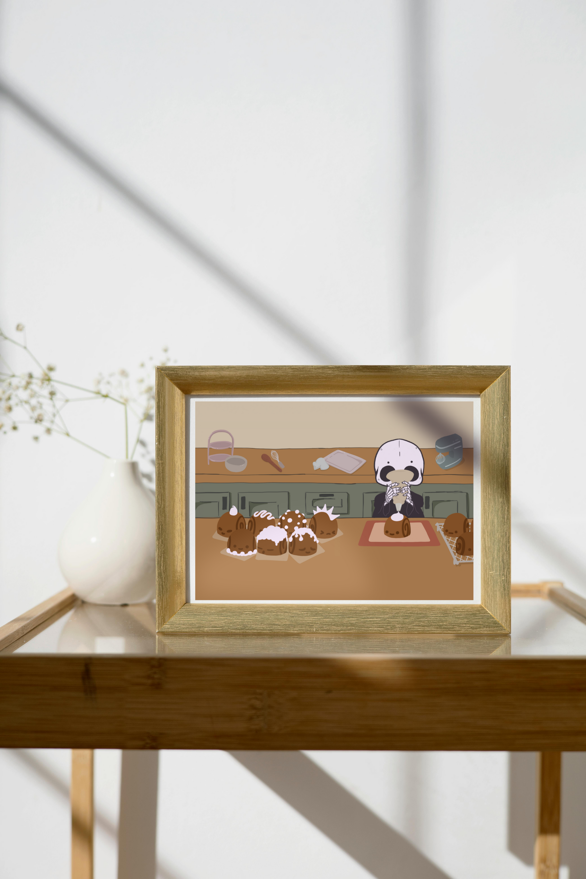 Framed art print on a glass and wooden side table next to a small plant in a white, ceramic vase. The framed art print is of an illustration of a skeleton character frosting cinnamon rolls that are personified with various facial expressions and frosting designs. The skeleton stands between two wooden counters. The background consists of various kitchen supplies including a stand mixer, whisk, baking sheet, bowl, green cabinets, and a beige wall. 