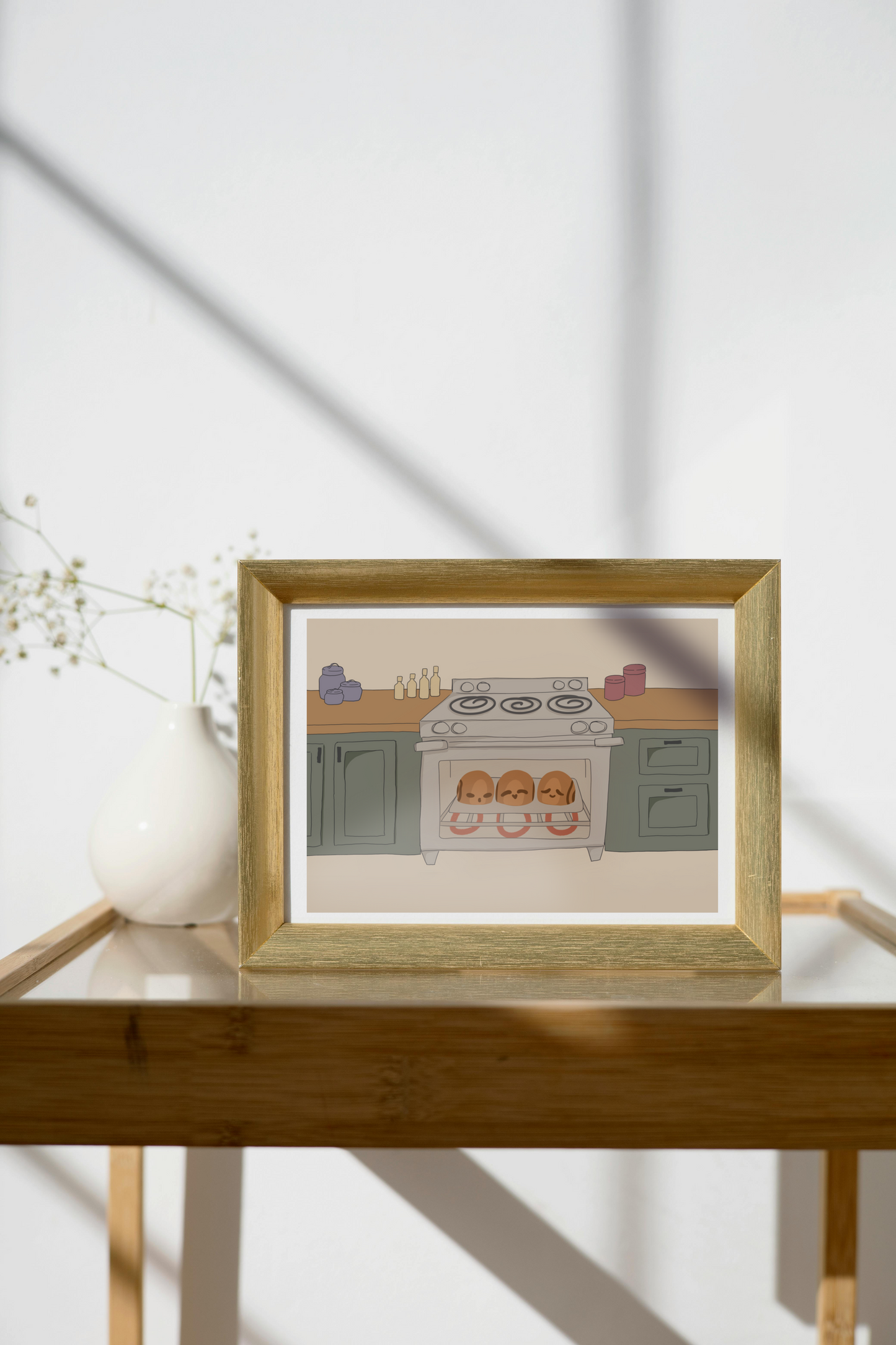 Framed art print on a glass and wooden side table next to a small plant in a white, ceramic vase. The framed art print is of an illustration of three cinnamon rolls personified with various facial expressions in an oven surrounded by wooden kitchen counters above green cabinets in front of a beige background.