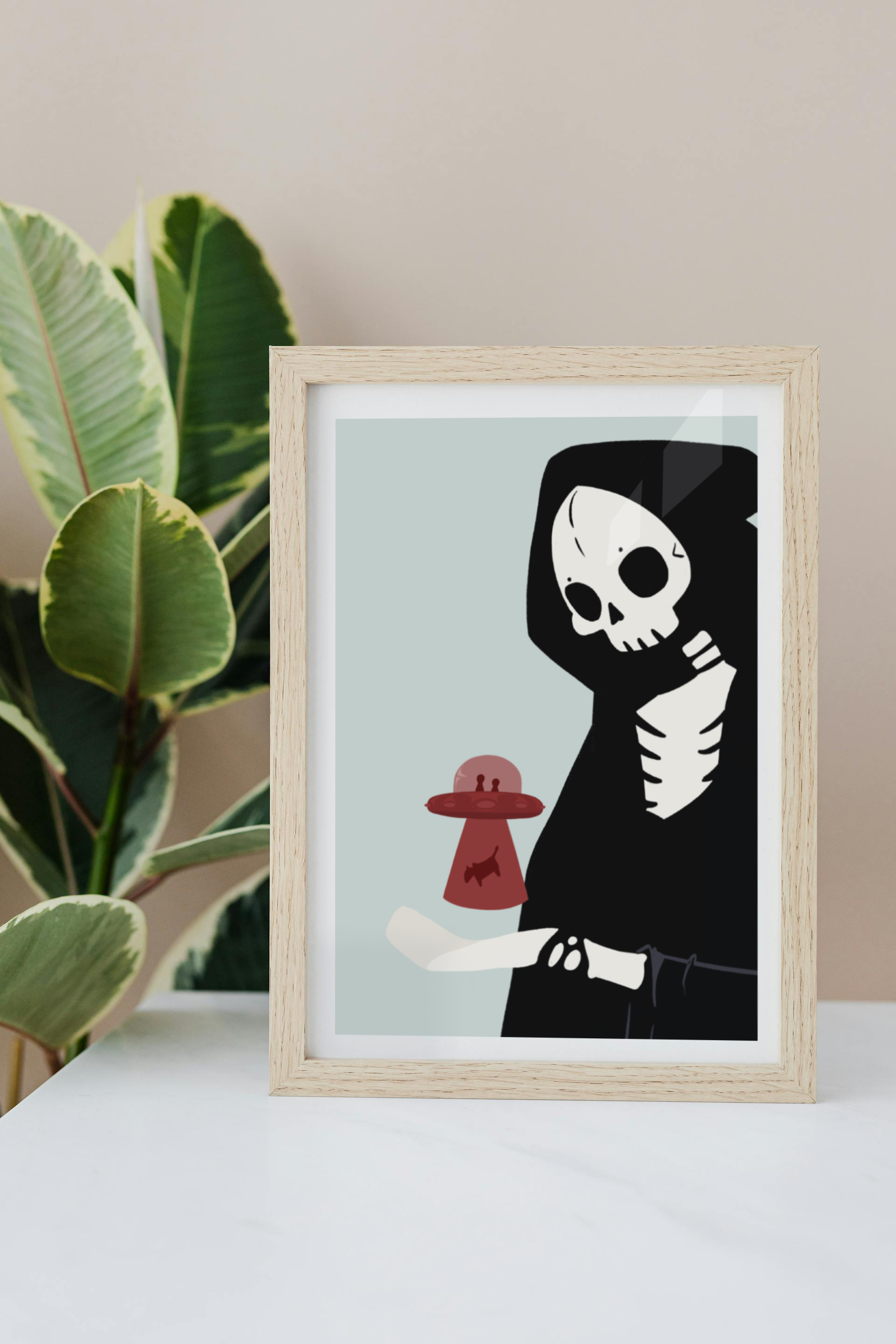 Framed art print on a white surface with a plant in the background of an illustration of a skeletal character with a red UFO abducting a cow hovering over its outreached hand in front of a light blue background.