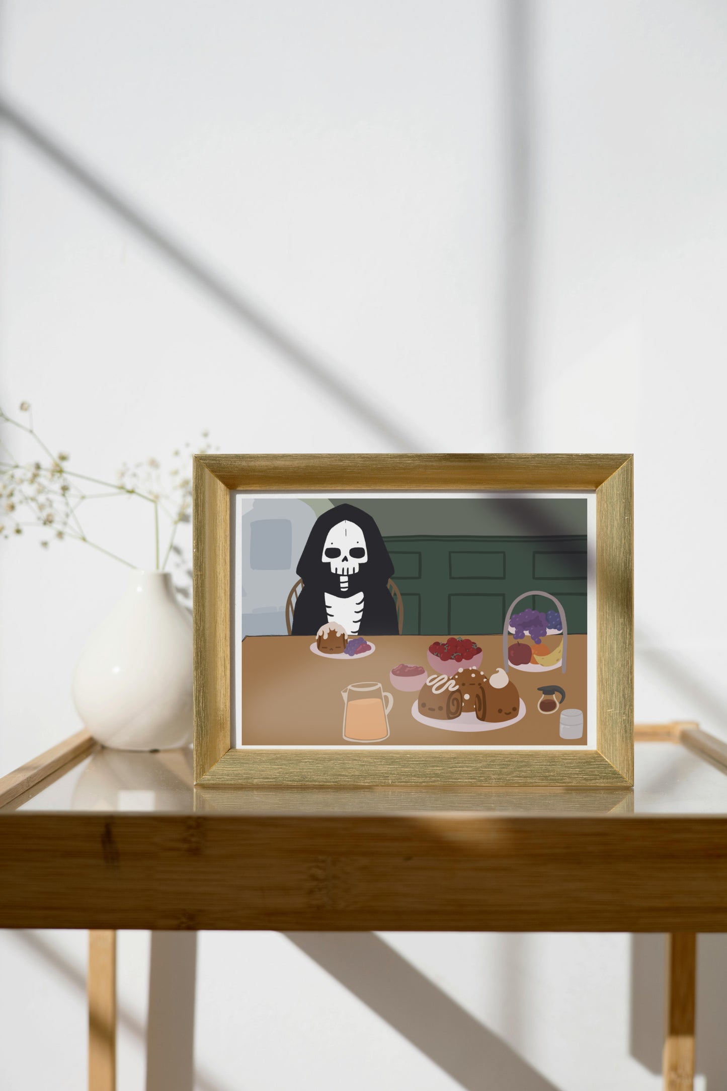 Framed art print on a glass and wooden side table next to a small plant in a white, ceramic vase. The framed art print is of an illustration of a skeleton character sitting at a table with brunch foods, including orange juice, cinnamon rolls personified with faces and various frosting designs, various fruits, a bowl of cherry tomatoes, syrup, jam, and powdered sugar. The background is of a green wall and hallway.