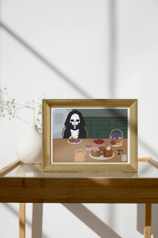 Framed art print on a glass and wooden side table next to a small plant in a white, ceramic vase. The framed art print is of an illustration of a skeleton character sitting at a table with brunch foods, including orange juice, cinnamon rolls personified with faces and various frosting designs, various fruits, a bowl of cherry tomatoes, syrup, jam, and powdered sugar. The background is of a green wall and hallway.