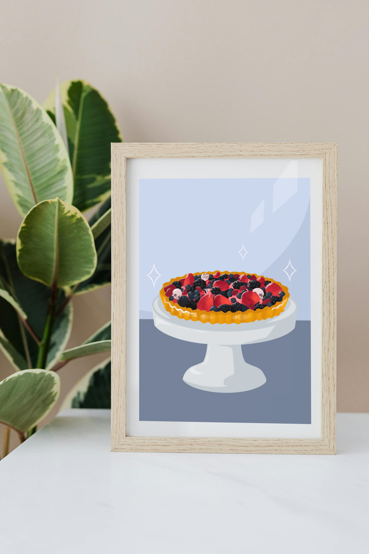 Framed art print on a white surface with a plant in the background of a fruit tart filled with strawberries, blackberries, raspberries, blueberries, tiny skulls, and tiny hand bones on a gray cake stand against a light blue and dark blue background. Three sparkles surround the fruit tart.