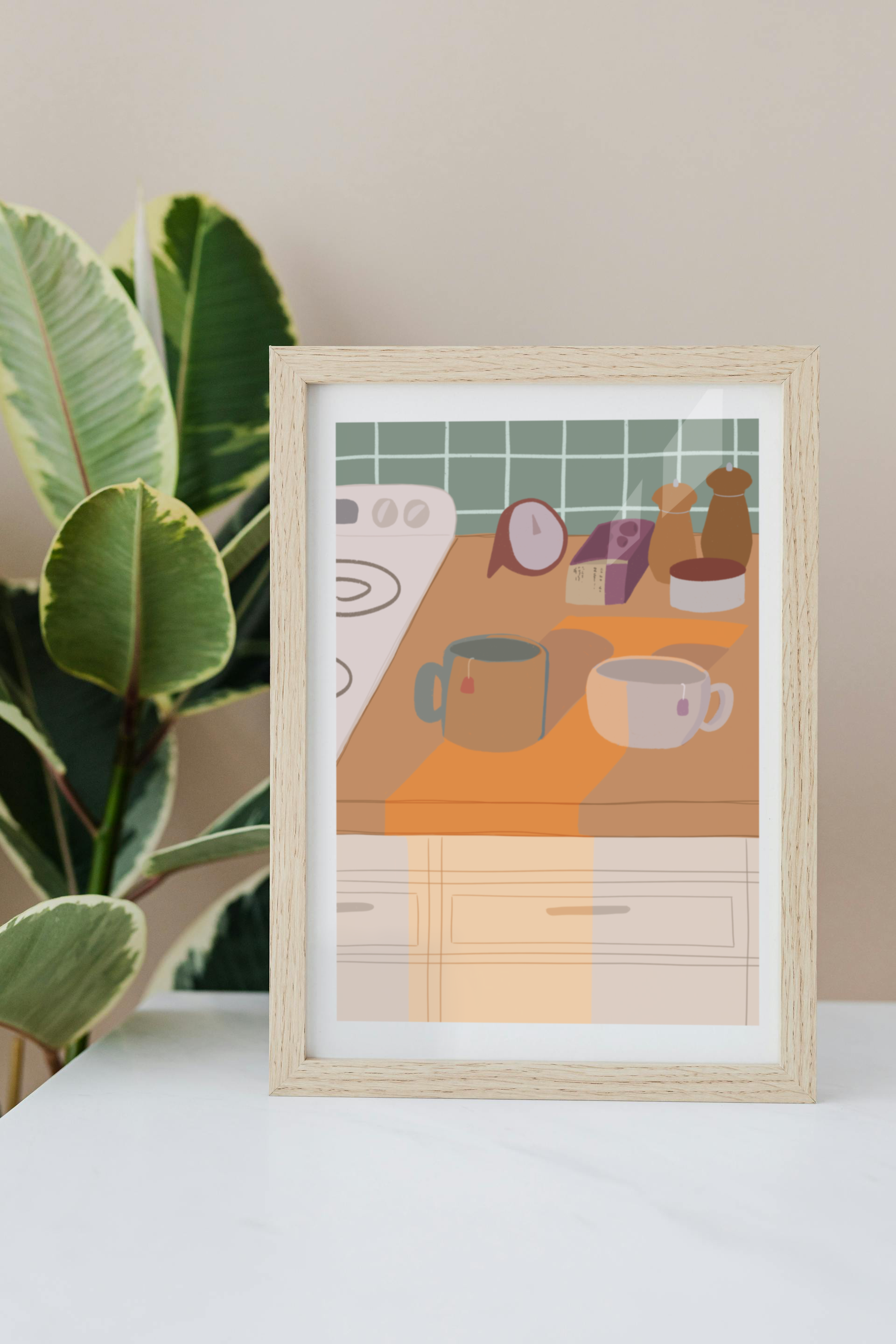 Framed art print on a white surface with a plant in the background of an illustration of a kitchen counter with two mugs holding tea bags in warm afternoon lighting. In the background are two pepper grinders, a tea box, a salt dish, and a timer. On the left is a white stovetop, white cabinet drawers at the bottom, and green tiles for the backsplash.