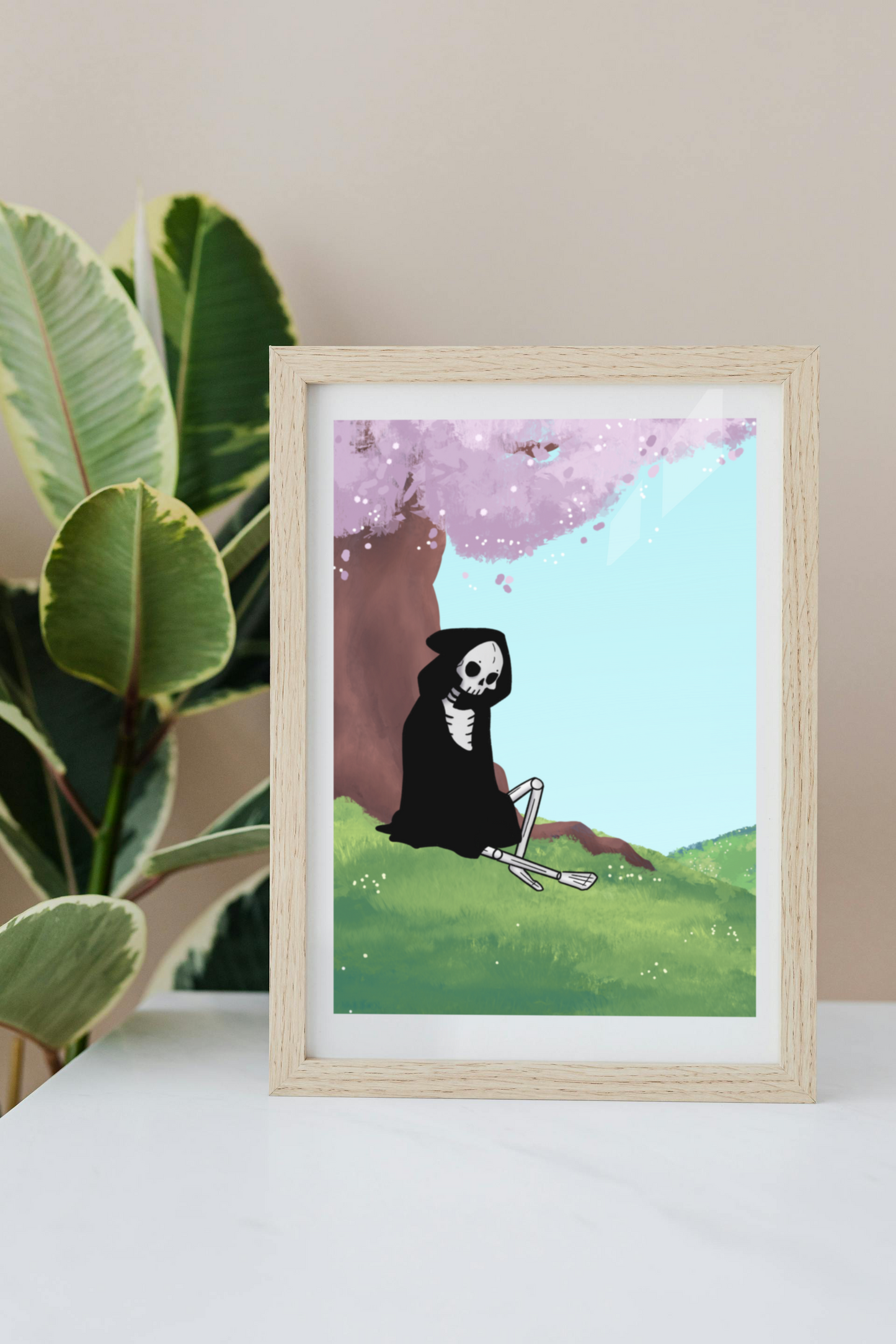 Framed artwork on a white surface with a plant in the background of a skeleton sitting under a cherry blossom tree.