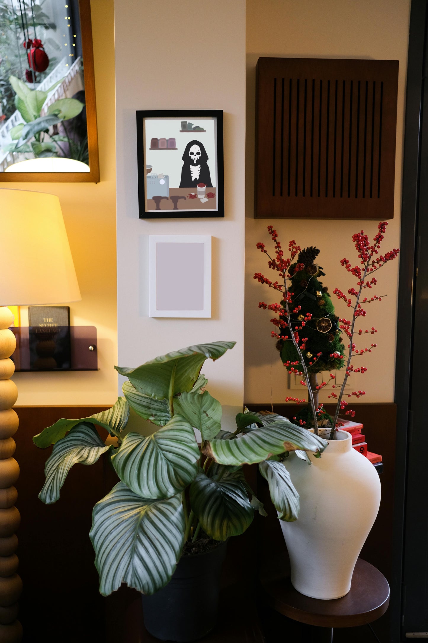 Framed art print hanging on a wall above a blank frame amongst house plants, a lamp, and wall decor. The framed art print is of an illustration of a skeleton character standing behind a wooden counter with an espresso machine, a to-go coffee cup, and 3 red imps atop it.  In the forefront are the tops of syrup bottles, in the background are 2 shelves holding coffee bags on a light blue wall.