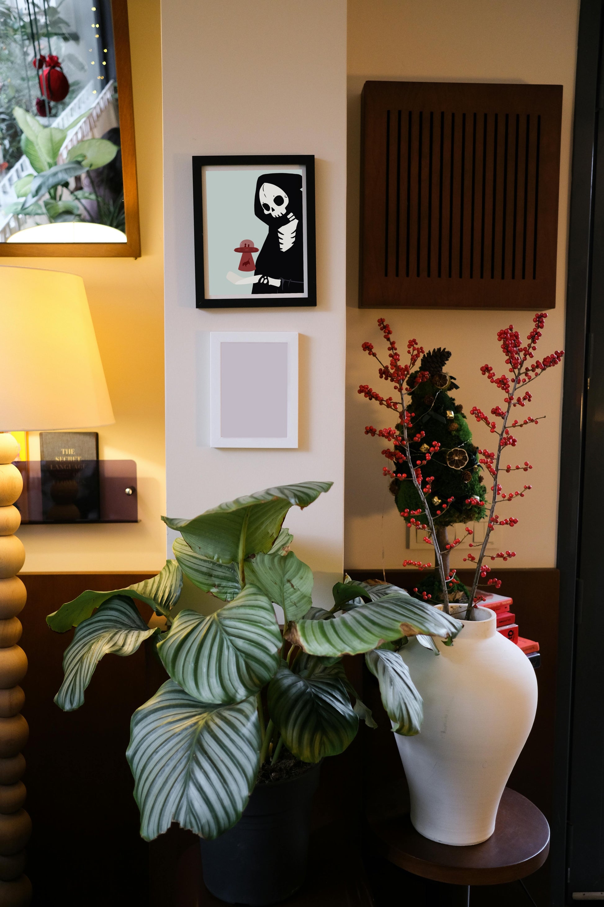 Framed art print hanging on a wall above a blank frame amongst house plants, a lamp, and wall decor. The framed art print is of an illustration of a skeletal character with a red UFO abducting a cow hovering over its outreached hand in front of a light blue background.
