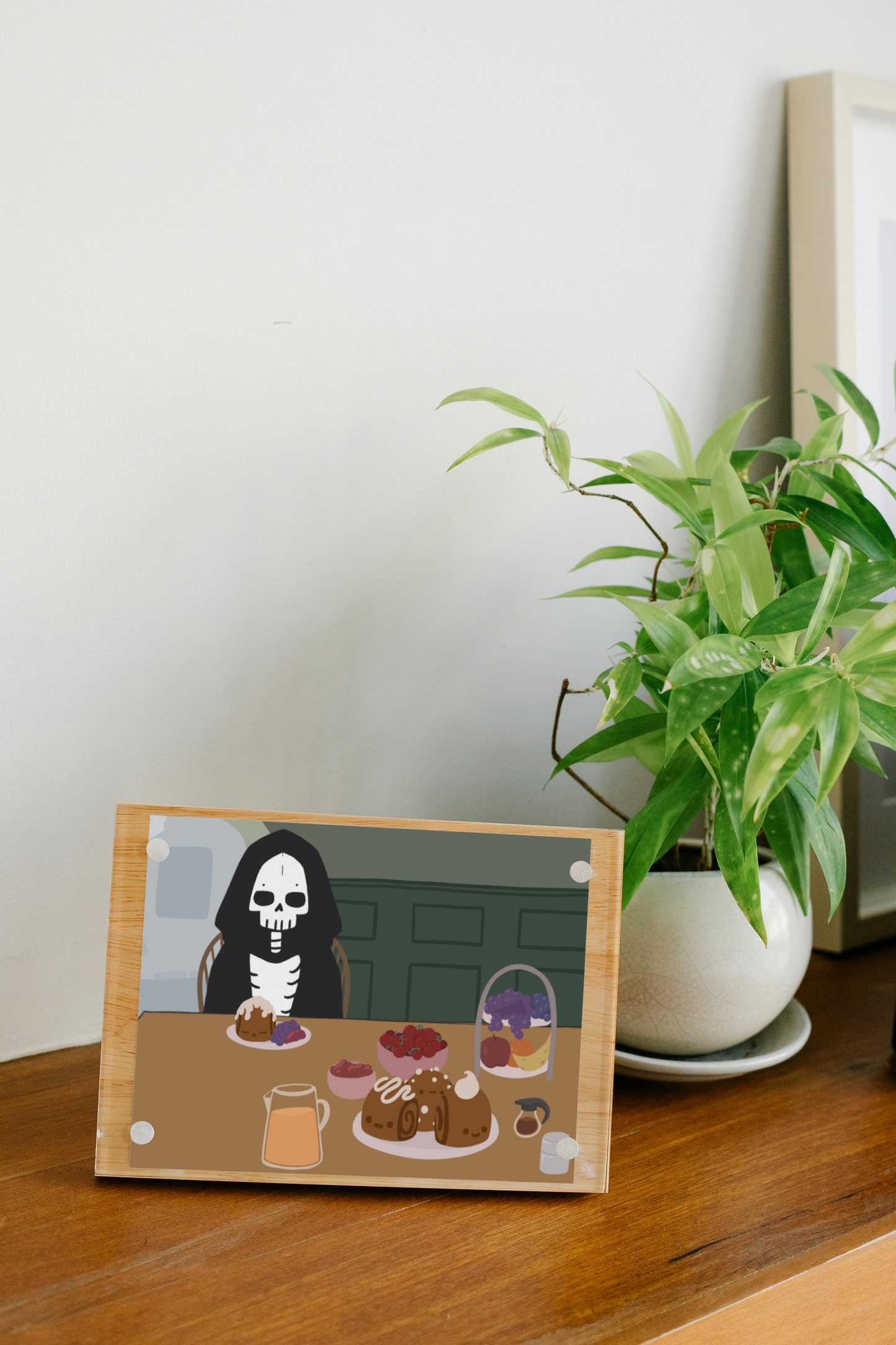 Framed art print on a wooden surface next to a small house plant of an iIllustration of a skeleton character sitting at a table with brunch foods, including orange juice, cinnamon rolls personified with faces and various frosting designs, various fruits, a bowl of cherry tomatoes, syrup, jam, and powdered sugar. The background is of a green wall and hallway.