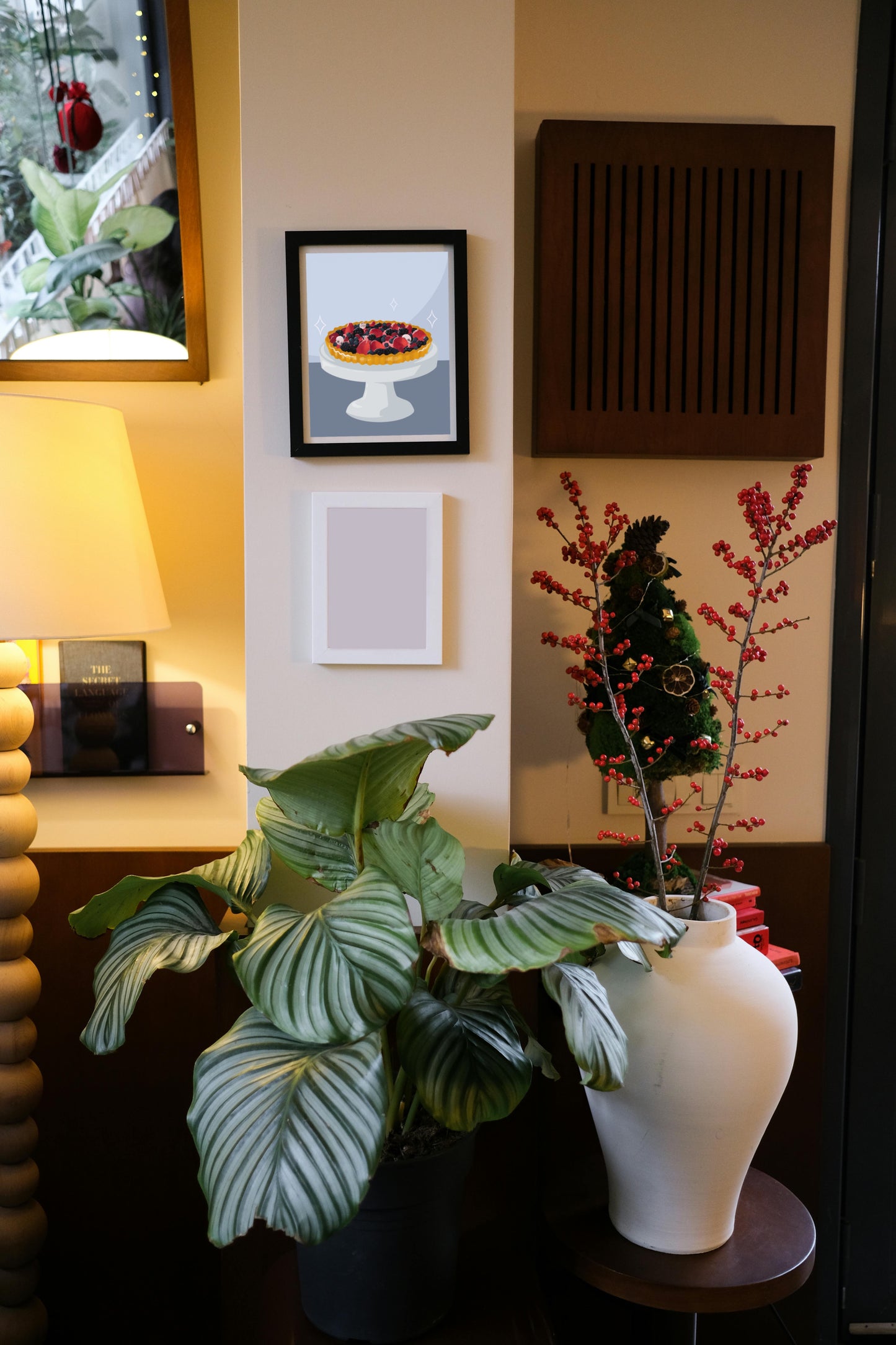 Framed art print hanging on a wall above a blank frame amongst house plants, a lamp, and wall decor. The framed art print is of an illustration of a fruit tart filled with strawberries, blackberries, raspberries, blueberries, tiny skulls, and tiny hand bones on a gray cake stand against a light blue and dark blue background. Three sparkles surround the fruit tart.
