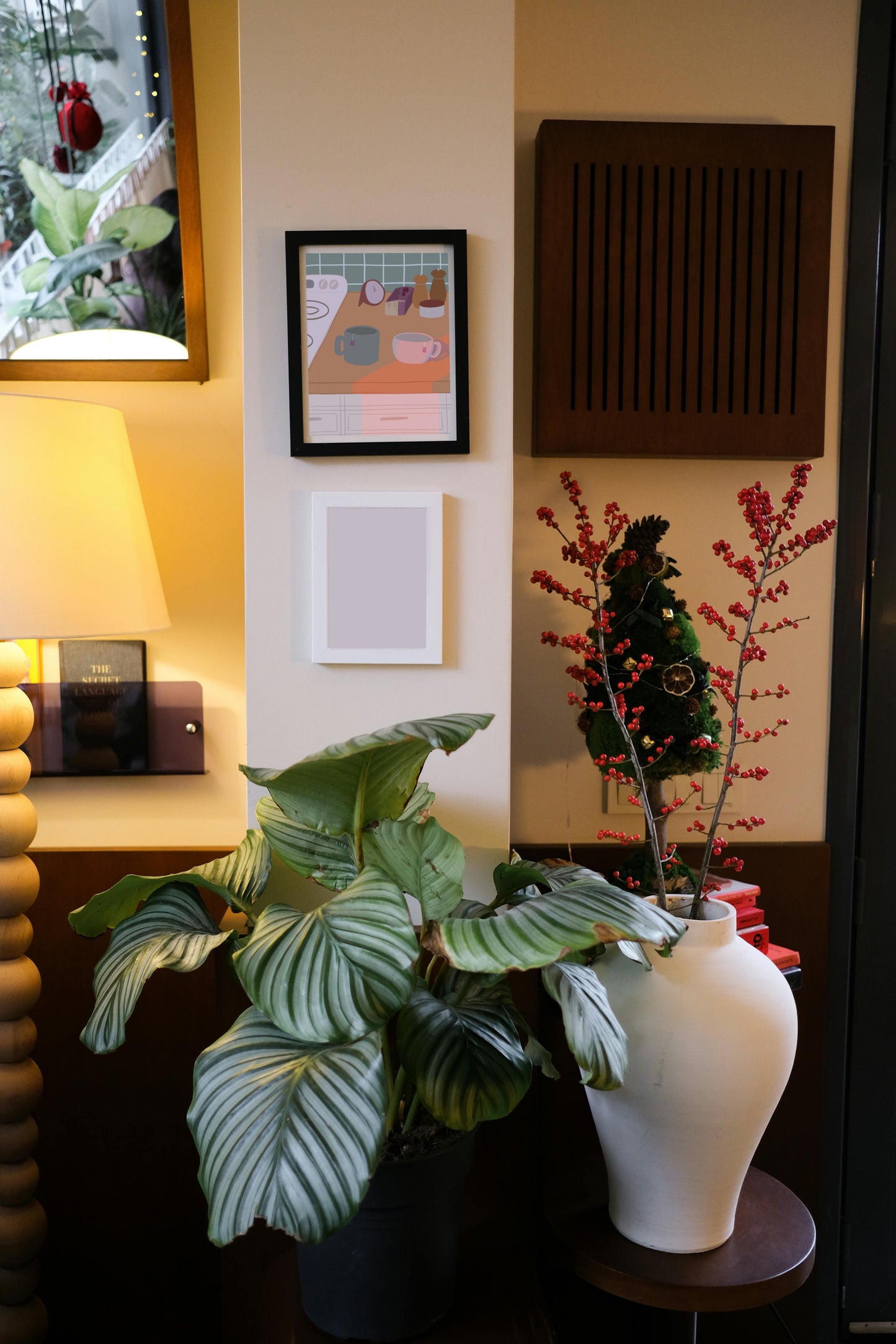 Framed art print hanging on a wall above a blank frame amongst house plants, a lamp, and wall decor. The framed art print is of an illustration of a kitchen counter with two mugs holding tea bags in cool evening lighting. In the background are two pepper grinders, a tea box, a salt dish, and a timer. On the left is a white stovetop, white cabinet drawers at the bottom, and green tiles for the backsplash.