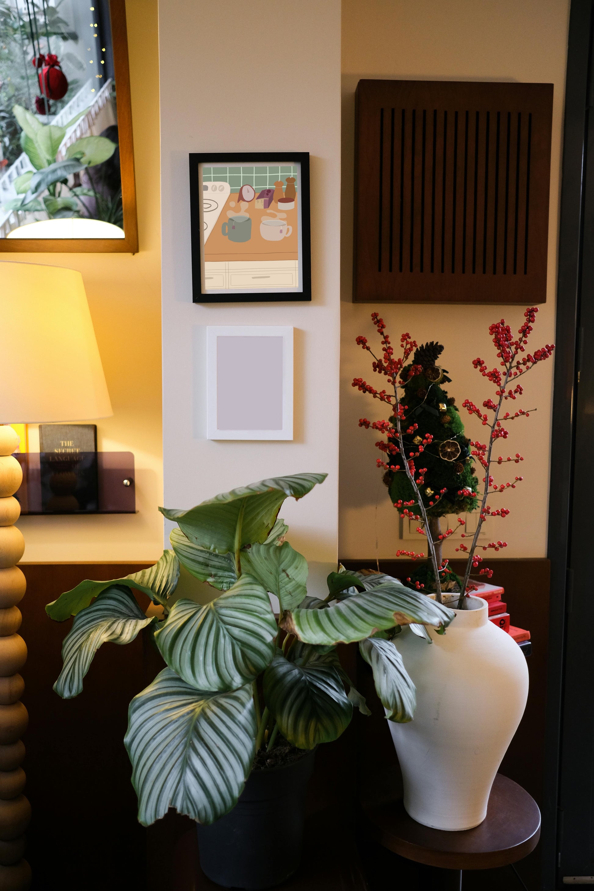 Framed art print hanging on a wall above a blank frame amongst house plants, a lamp, and wall decor. The framed art print is of an illustration of a kitchen counter with two steaming mugs holding tea bags in morning lighting. In the background are two pepper grinders, a tea box, a salt dish, and a timer. On the left is a white stovetop, white cabinet drawers at the bottom, and green tiles for the backsplash.