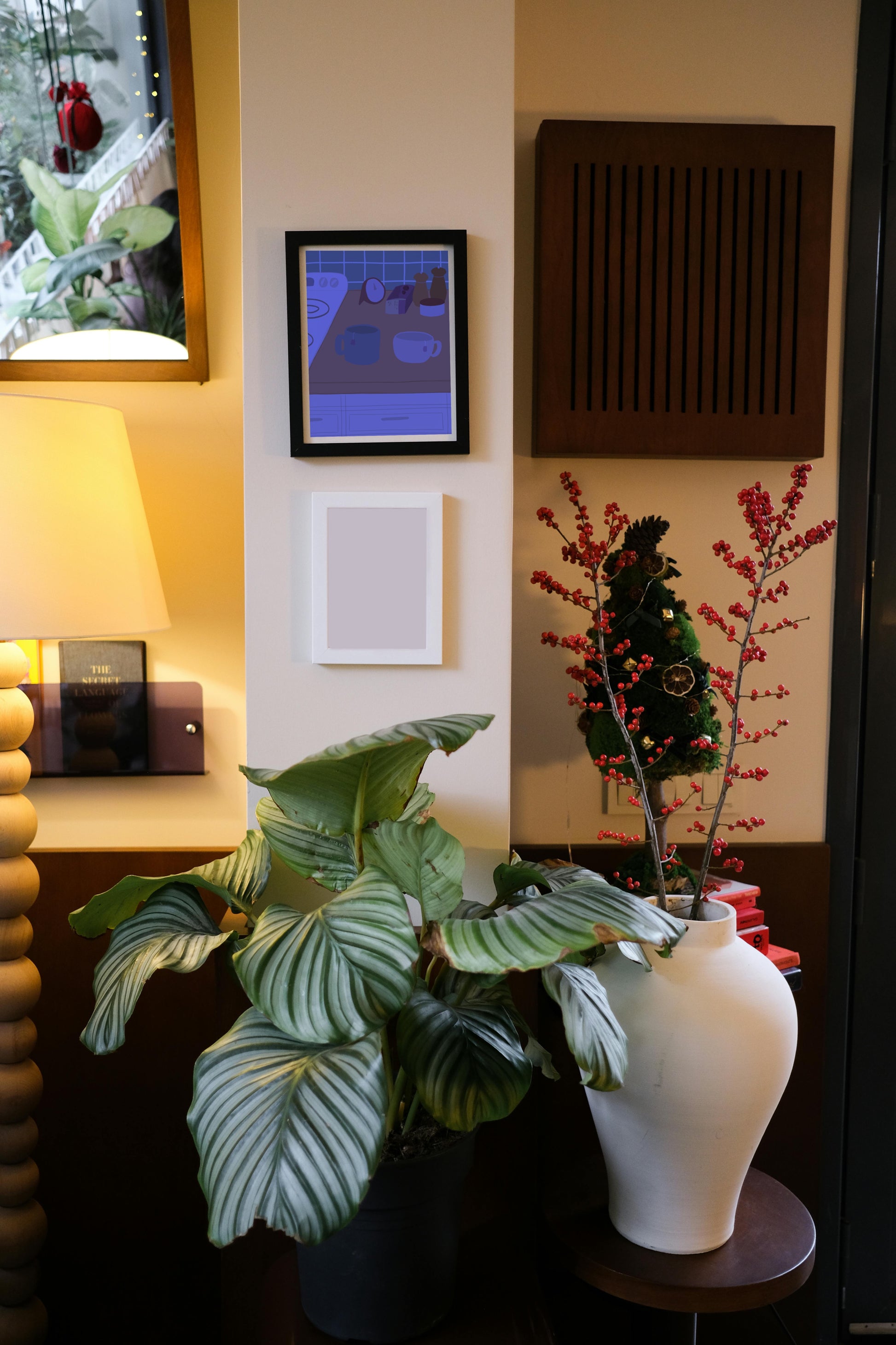 Framed art print hanging on a wall above a blank frame amongst house plants, a lamp, and wall decor. The framed art print is of an illustration of a kitchen counter with two mugs holding tea bags in blue night lighting. In the background are two pepper grinders, a tea box, a salt dish, and a timer. On the left is a white stovetop, white cabinet drawers at the bottom, and green tiles for the backsplash.