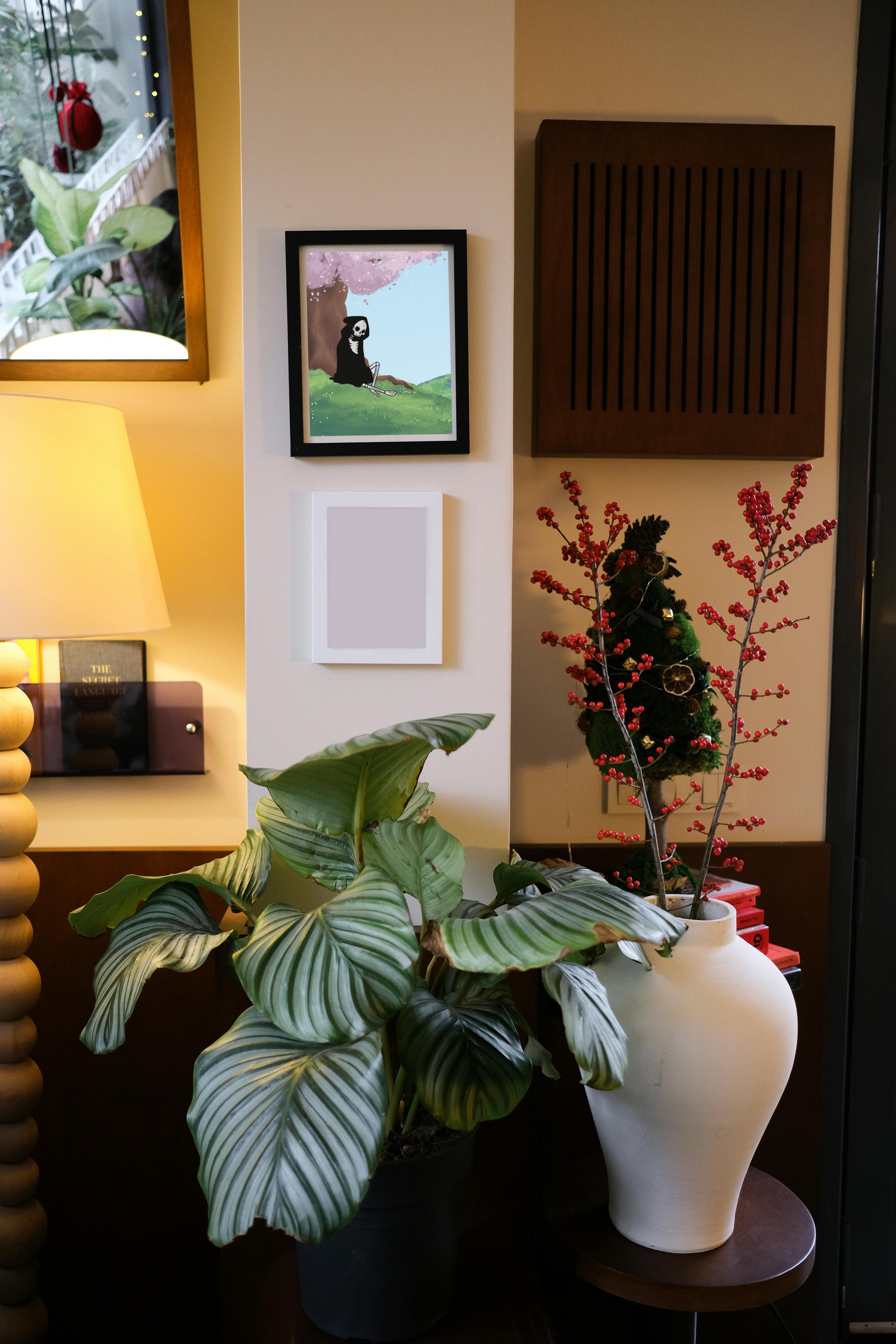 Framed art print hanging on a wall above a blank frame amongst house plants, a lamp, and wall decor. The framed art print is of a  skeleton under a cherry blossom tree with a light blue sky in a hilly field.