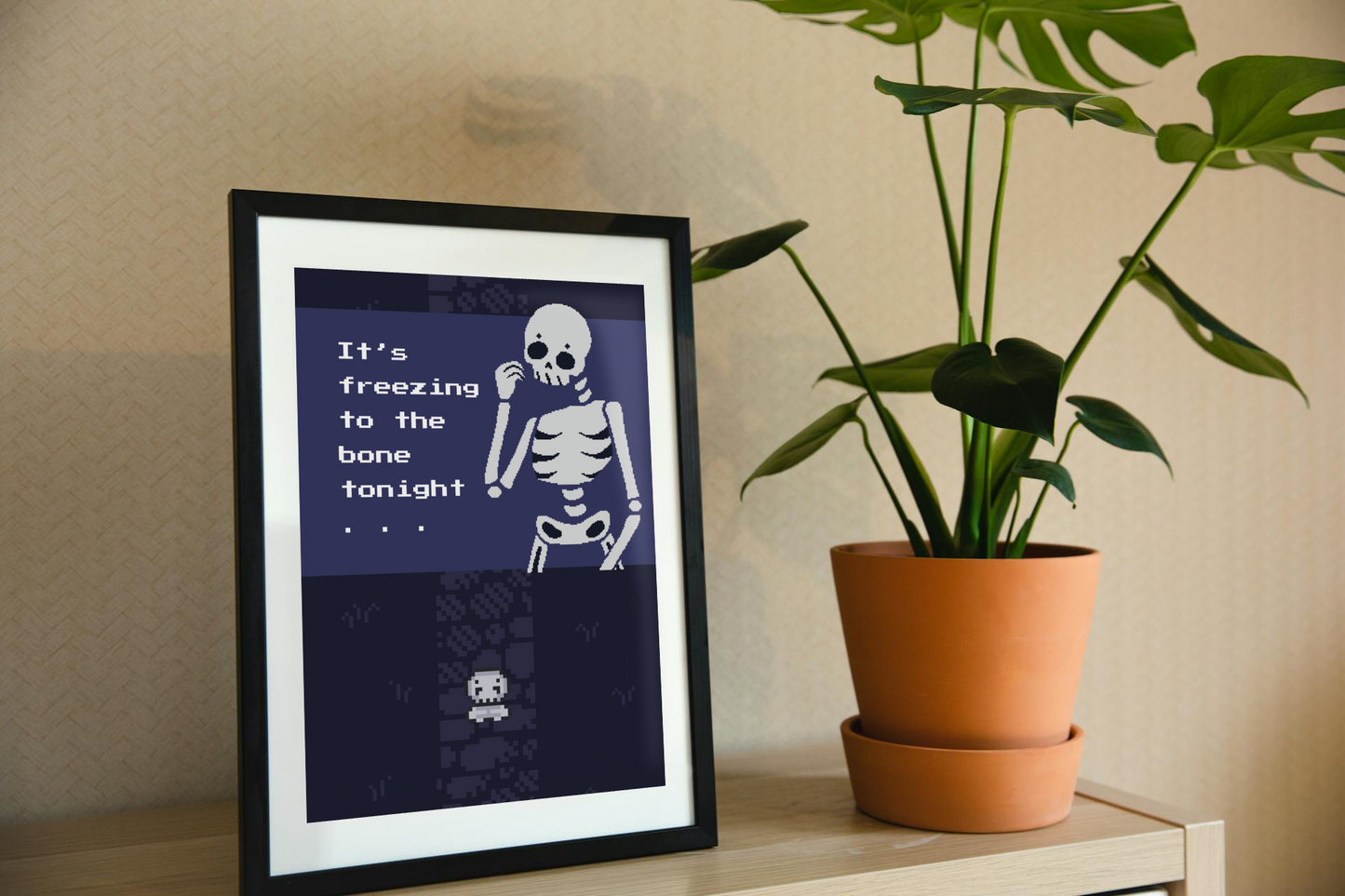 Framed art print on a shelf next to a house plant. The framed art print is of an illustration of a pixelated skeleton with text 'It's freezing to the bone tonight' on a dark blue background. Behind that is a pixelated dark purple path on pixelated dark blue grass, with a smaller pixelated skeleton on top of that.