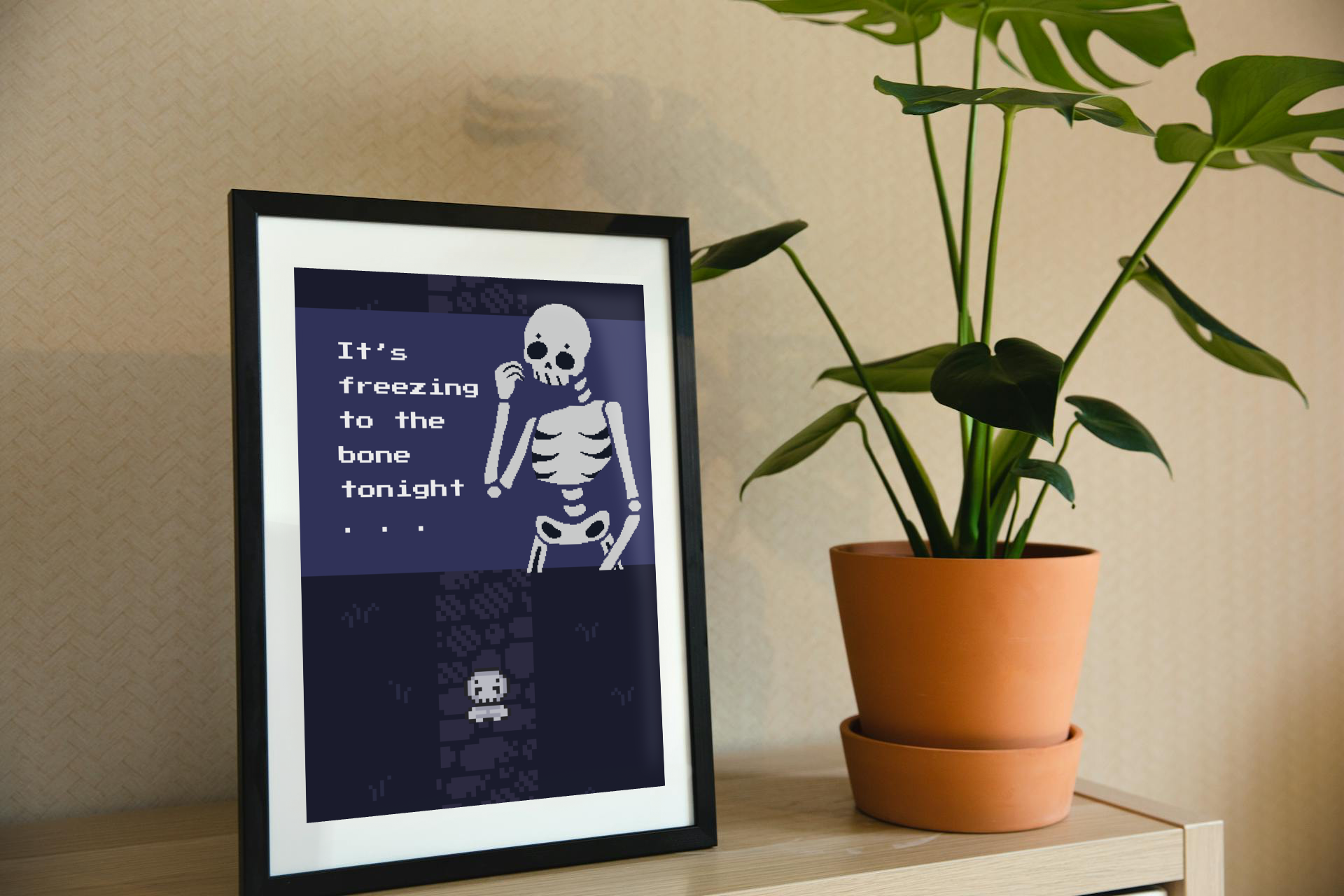 Framed art print on a shelf next to a house plant. The framed art print is of an illustration of a pixelated skeleton with text 'It's freezing to the bone tonight' on a dark blue background. Behind that is a pixelated dark purple path on pixelated dark blue grass, with a smaller pixelated skeleton on top of that.