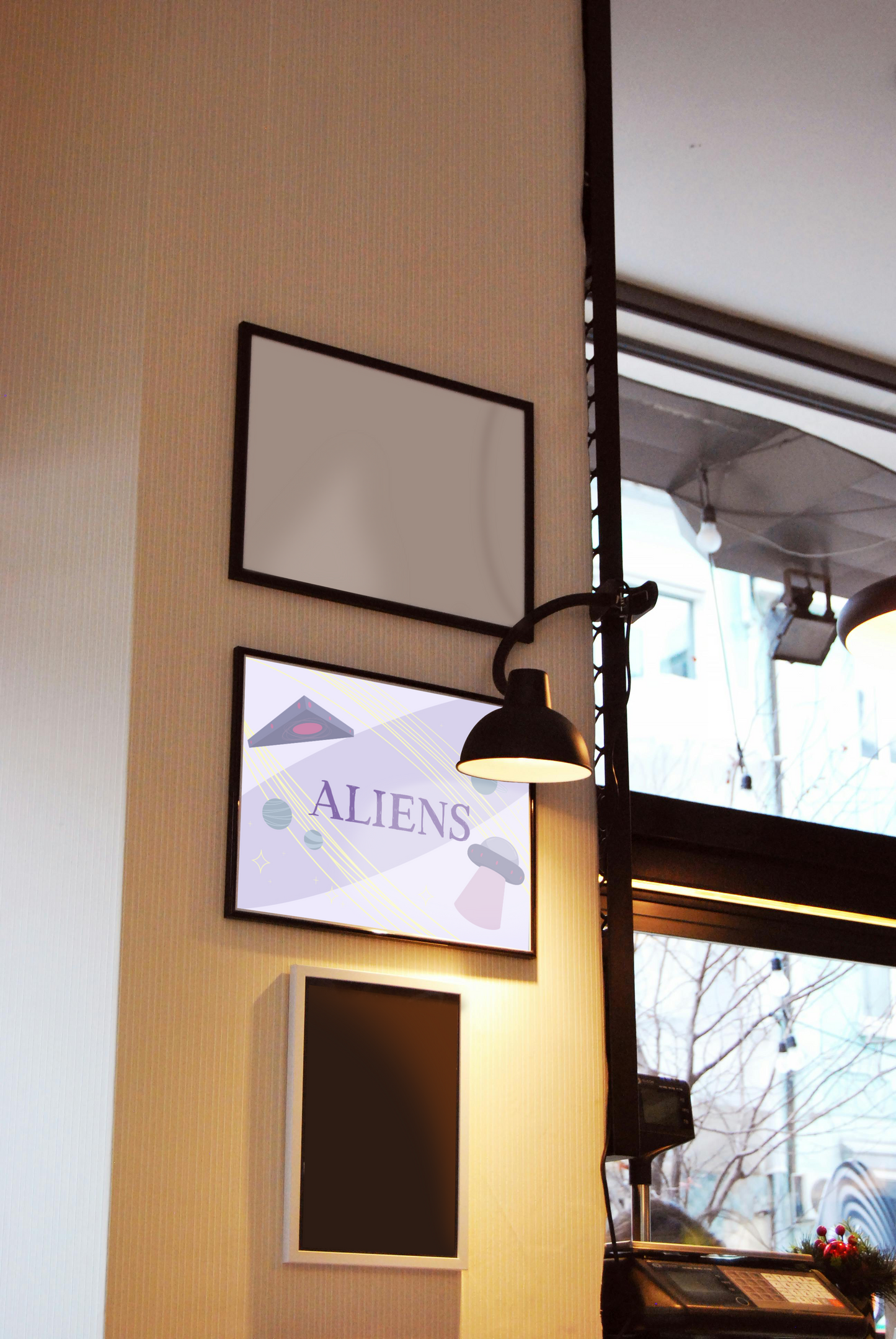 Framed art print hanging on a wall between blank frames next to a lit lamp and large window. The framed art print is of an illustration of a triangle UFO, a circular UFO, three green planets, and various yellow stars on a light purple background with the word 'ALIENS' in the center in dark purple.
