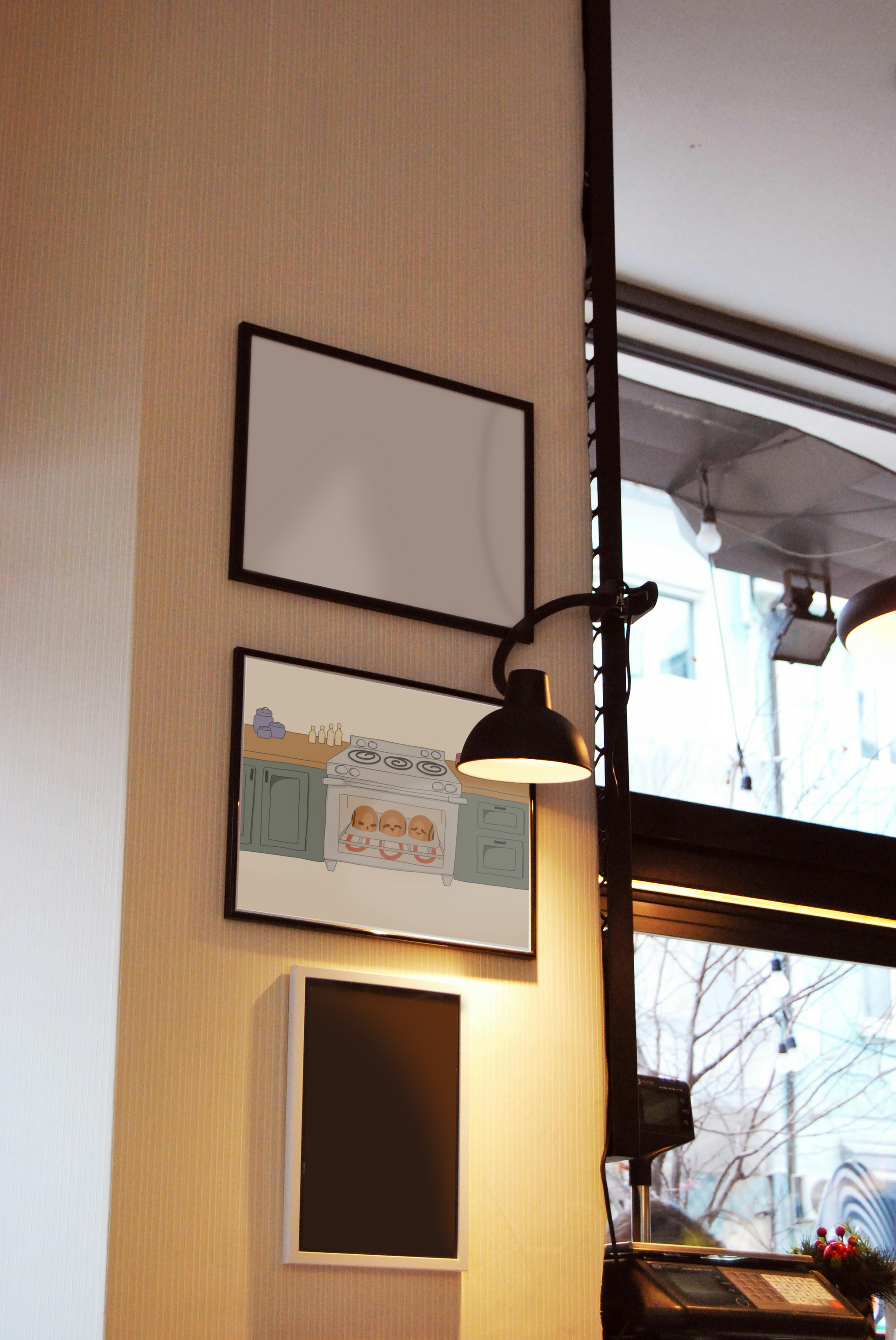 Framed art print hanging on a wall between blank frames next to a lit lamp and large window. The framed art print is of an illustration of three cinnamon rolls personified with various facial expressions in an oven surrounded by wooden kitchen counters above green cabinets in front of a beige background.